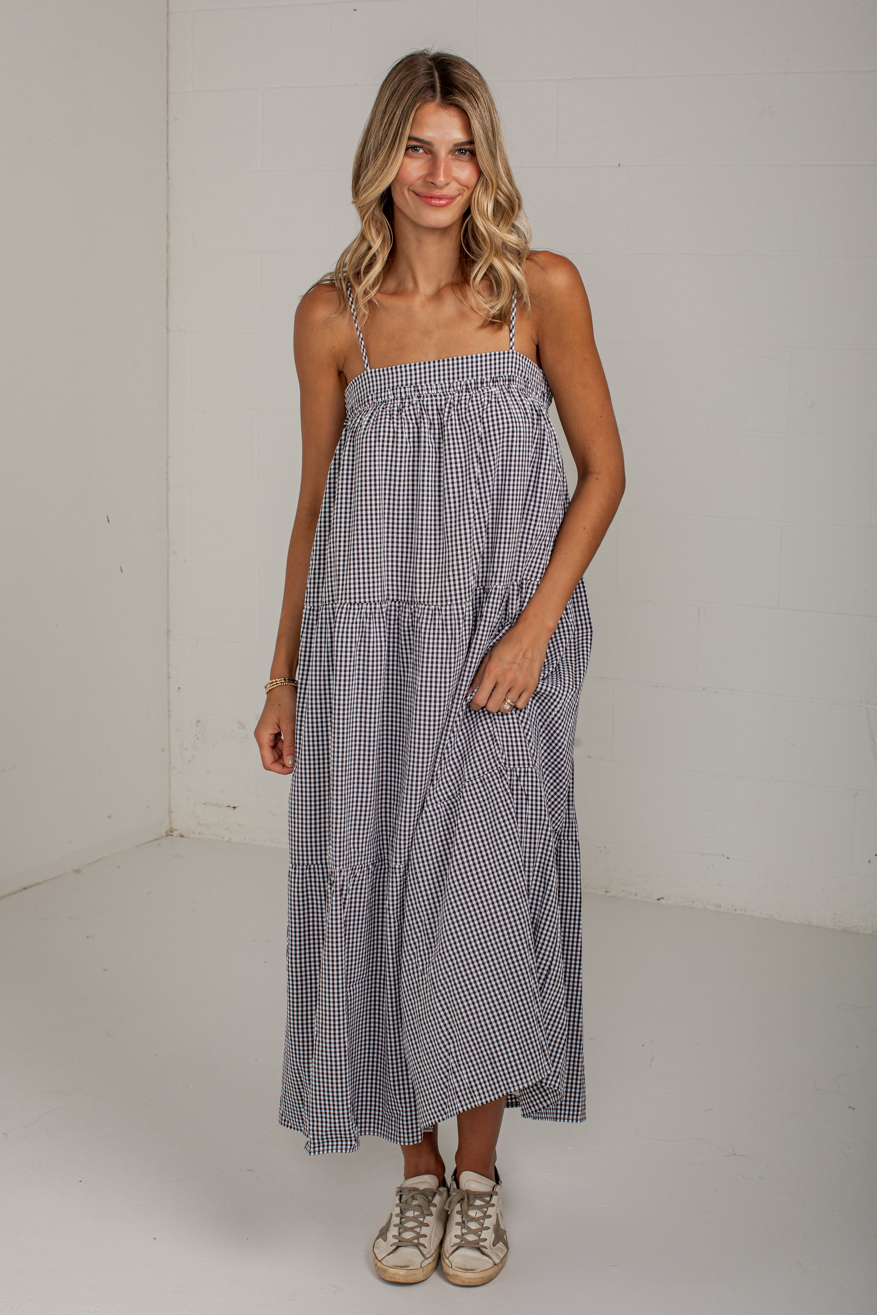 A woman modeling a sleeveless, tiered maxi dress with a small black and white gingham pattern, standing in a white studio corner.