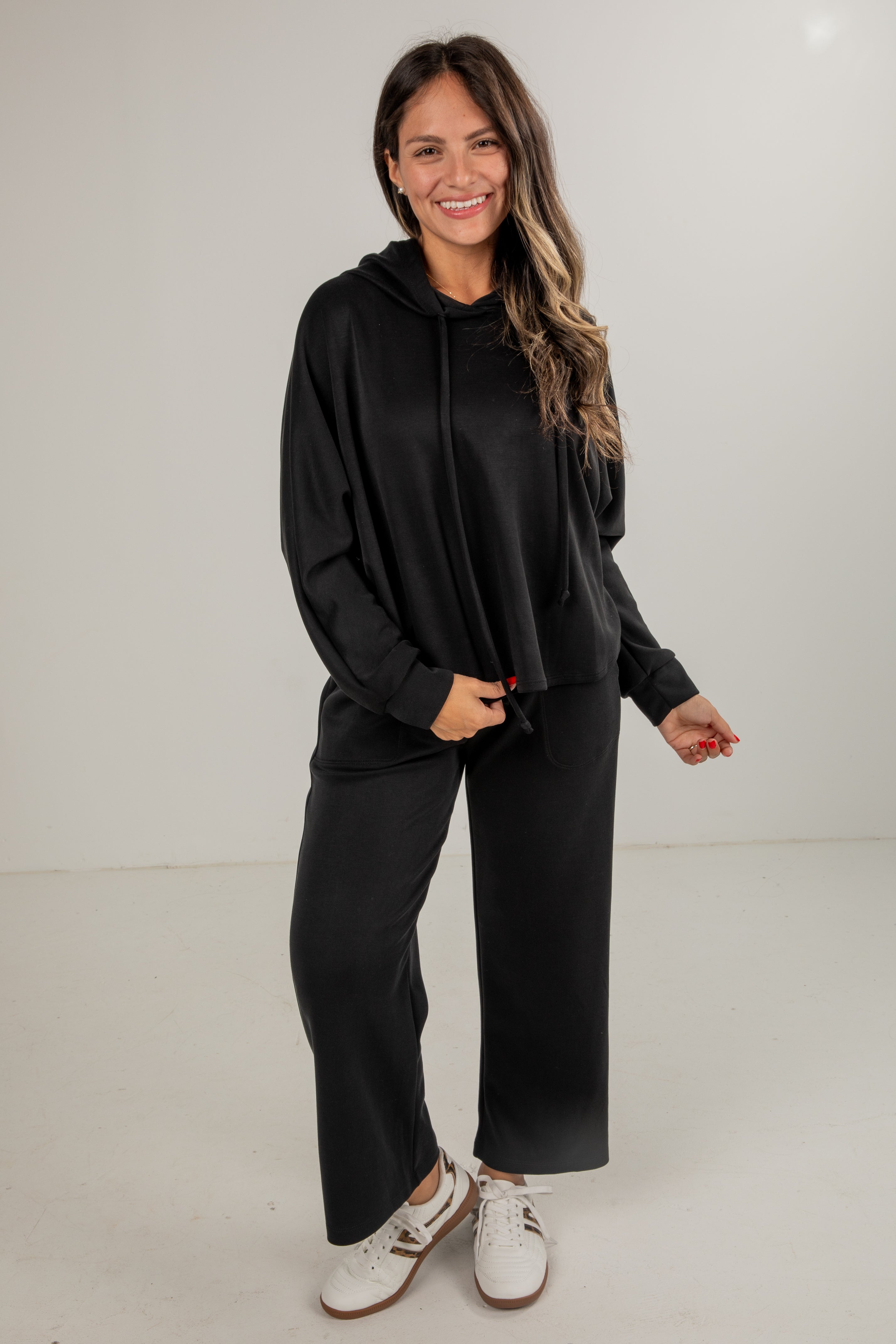 Woman wearing a black hoodie and pants with white sneakers on a plain background