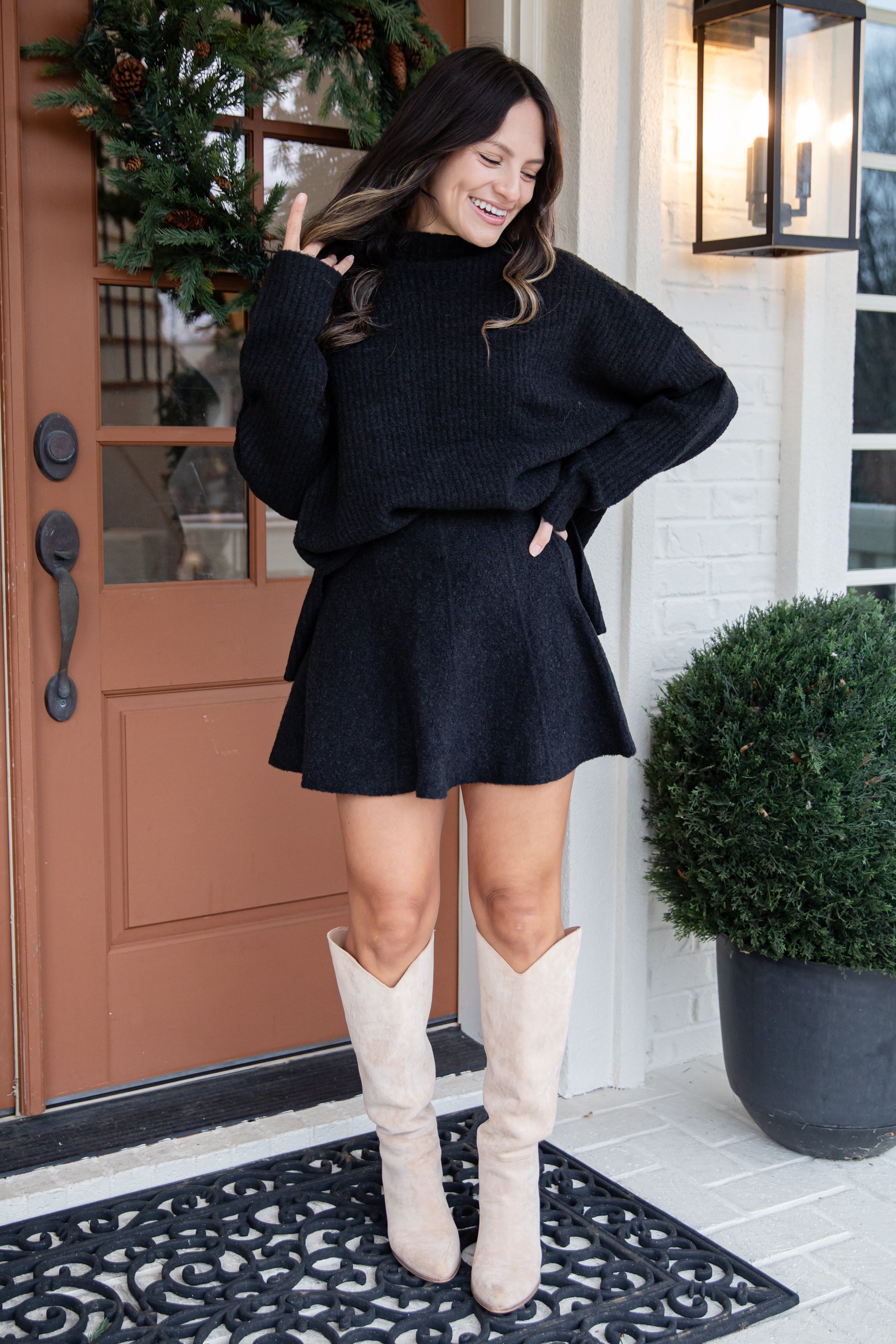 Woman in a black outfit with beige boots standing in front of a door.