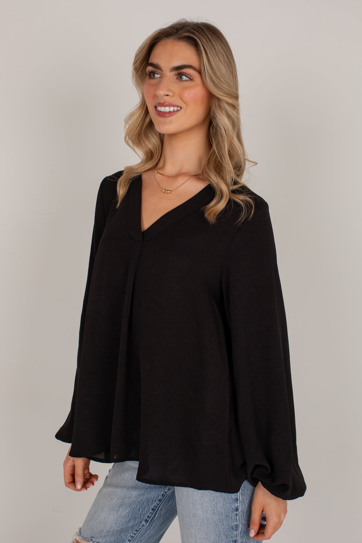 A woman modeling a black long-sleeve blouse with a V-neck and gathered cuffs, paired with light blue jeans, against a plain light gray studio background.