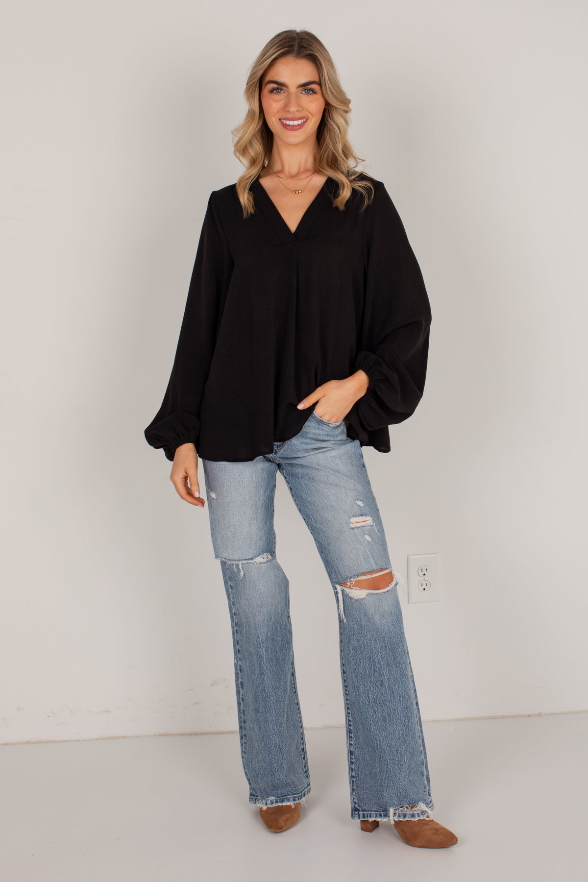 A woman modeling a black long-sleeve blouse with a V-neck and balloon sleeves, paired with light-wash distressed flare jeans and brown shoes, standing against a plain light gray wall.