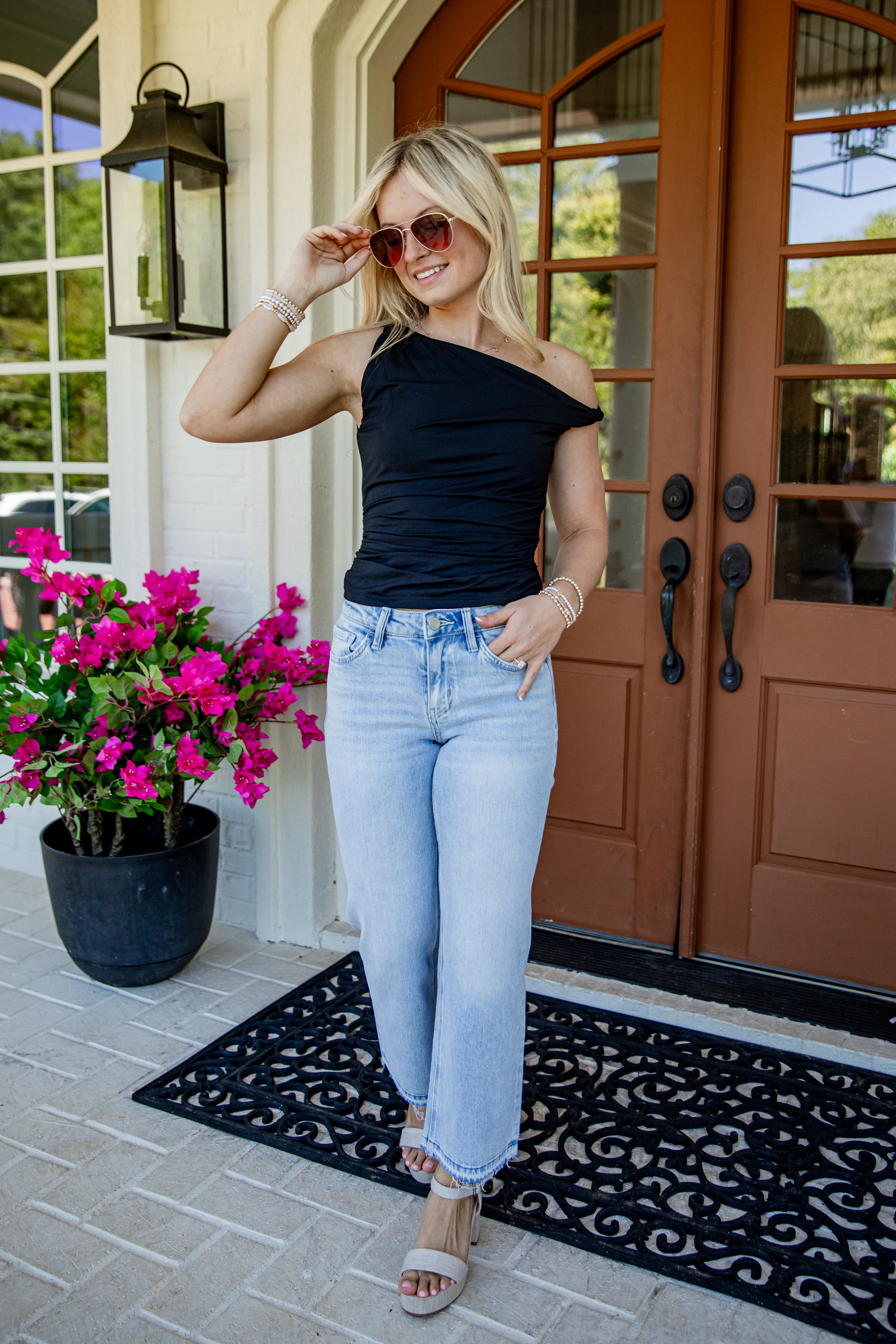 A woman stands on a front porch wearing a black off-the-shoulder top, light-wash blue jeans, and beige sandals. She is posing in front of brown double doors with a potted pink flower on the left.
