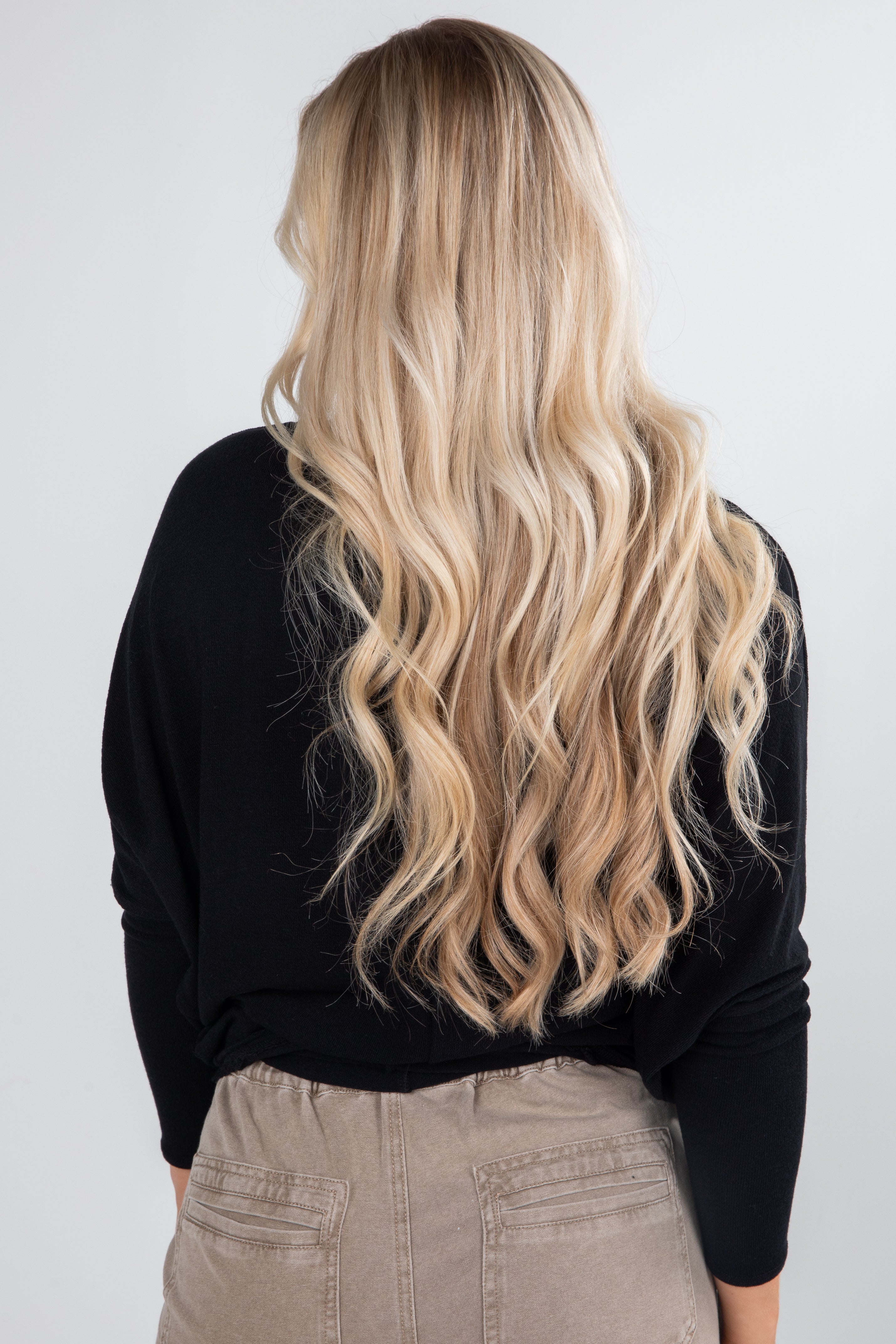 Person with long, wavy blonde hair wearing a black sweater and beige pants on a white background