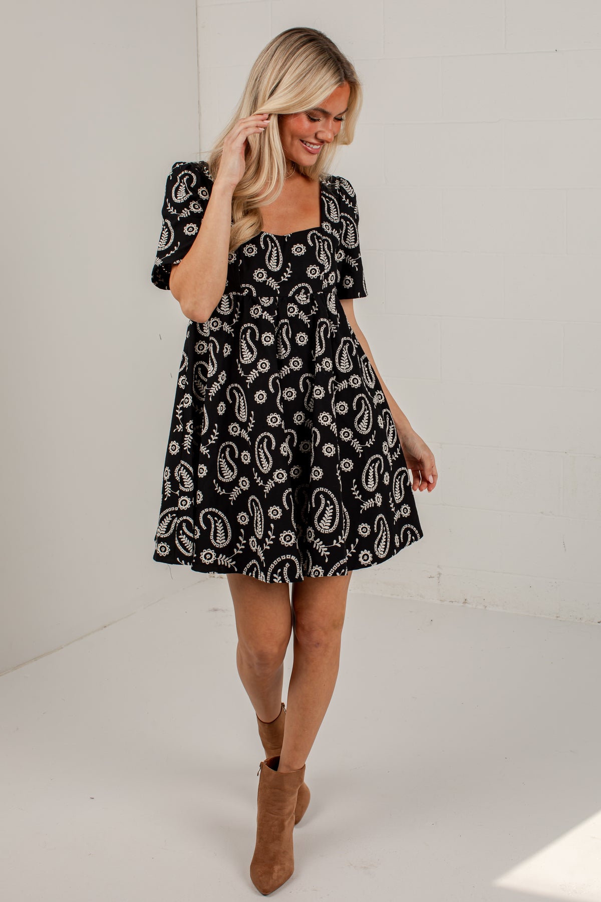 A woman modeling a short puff-sleeve dress with a black and white paisley pattern, paired with brown ankle boots, in a bright white studio setting.