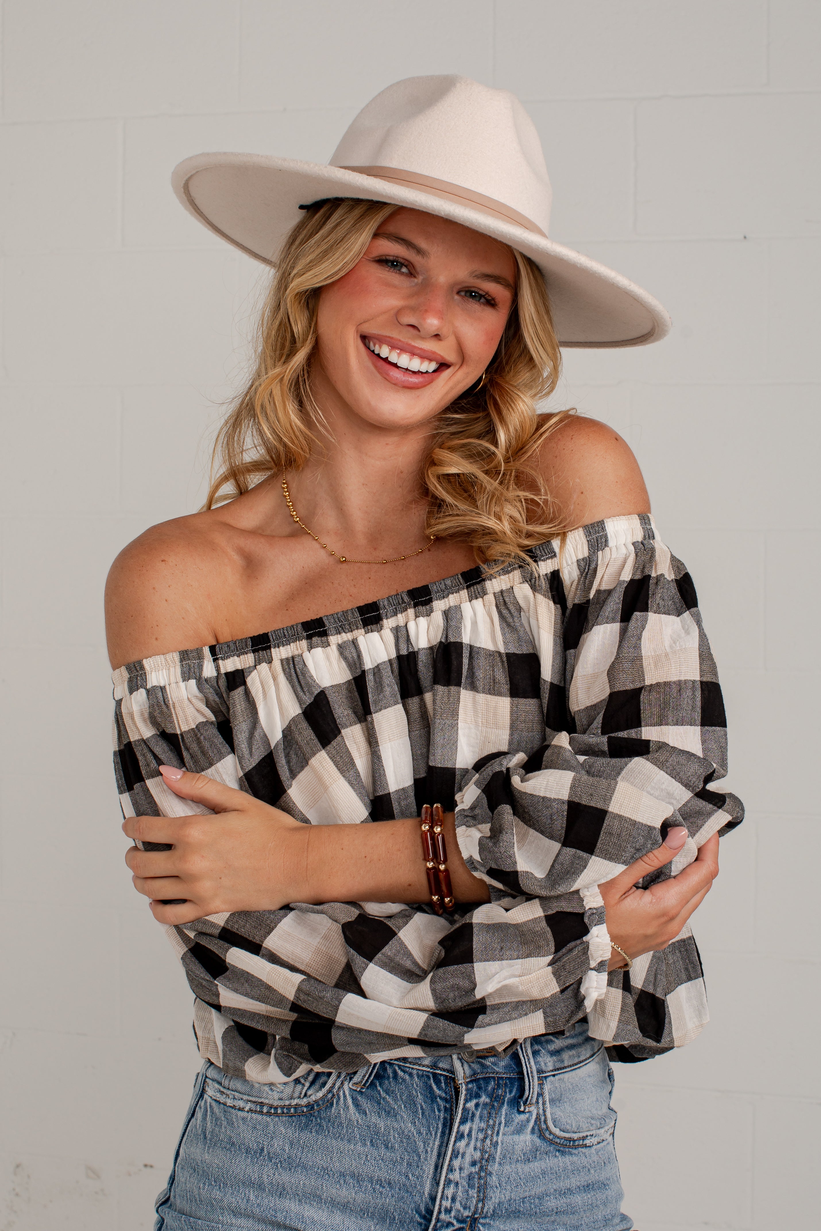 A smiling woman wearing an off-the-shoulder black and white buffalo plaid top, a cream wide-brim fedora hat, and blue jeans, posing against a light gray brick wall.