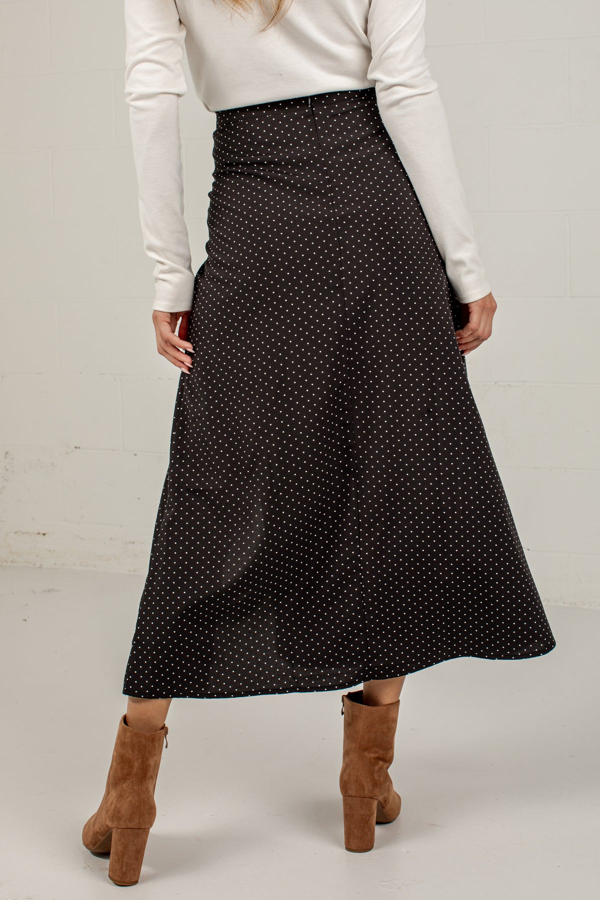A woman wearing a black midi skirt with a small white polka dot pattern, paired with a white long-sleeve top and brown suede block-heel ankle boots, shown from the back against a light gray studio background.