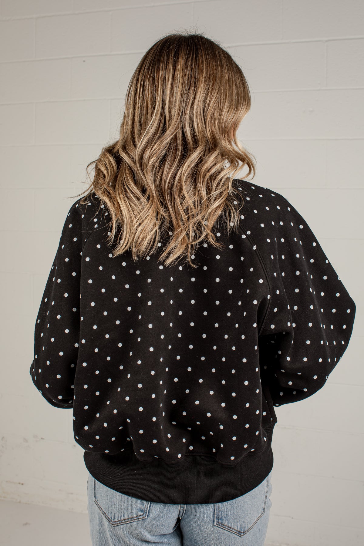 A woman viewed from behind wearing a black crewneck sweatshirt with white polka dots, paired with light blue jeans, standing against a white brick wall.
