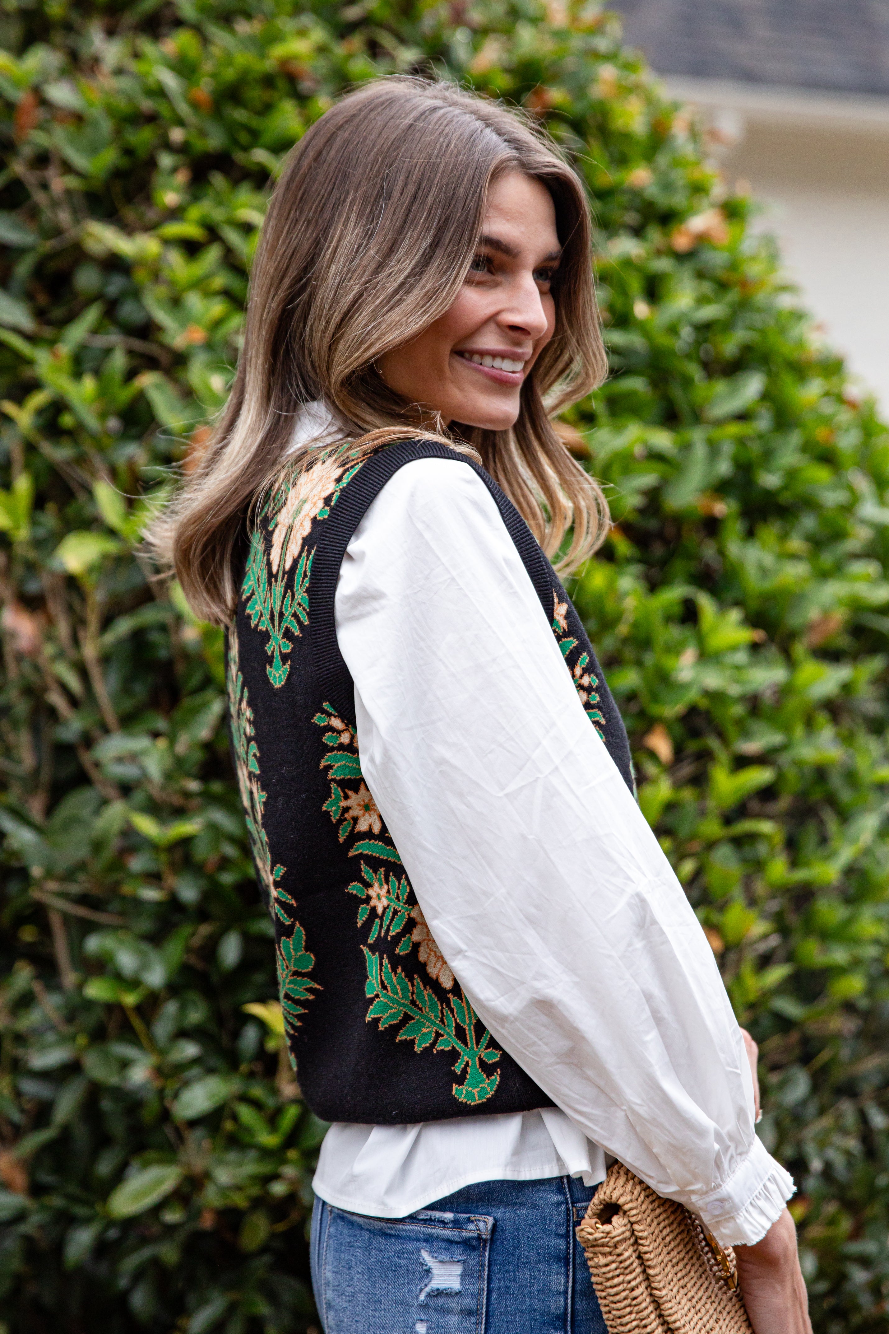 Woman wearing a black embroidered vest over a white blouse with blue jeans, standing in front of green bushes.