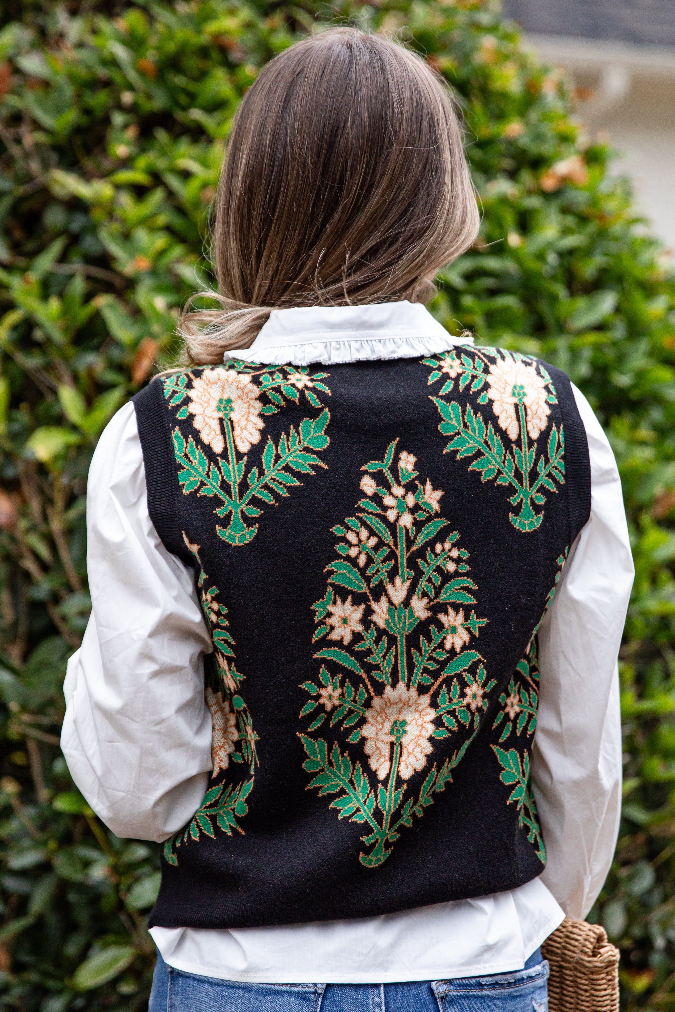 Person wearing a black embroidered vest over a white shirt with a green bush background