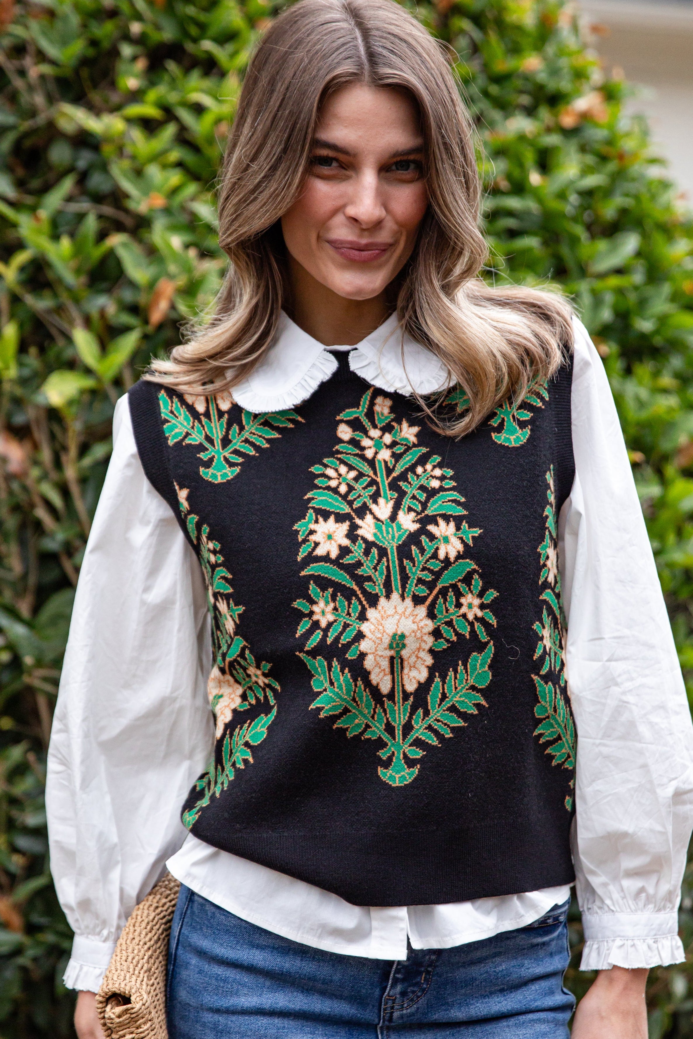 Woman wearing a black embroidered top with green and gold designs, standing in front of green foliage.