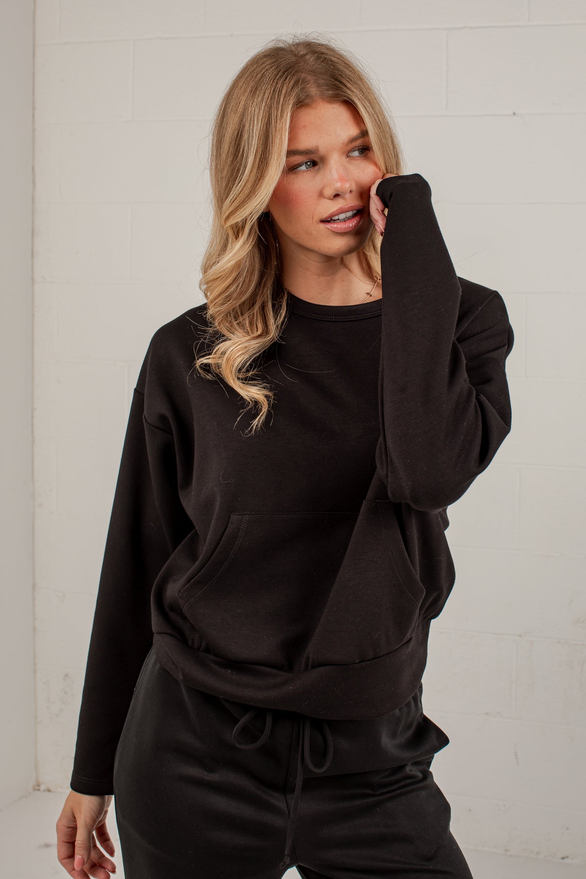 A woman modeling a black long-sleeve crewneck sweatshirt and matching black drawstring sweatpants against a white brick wall background.