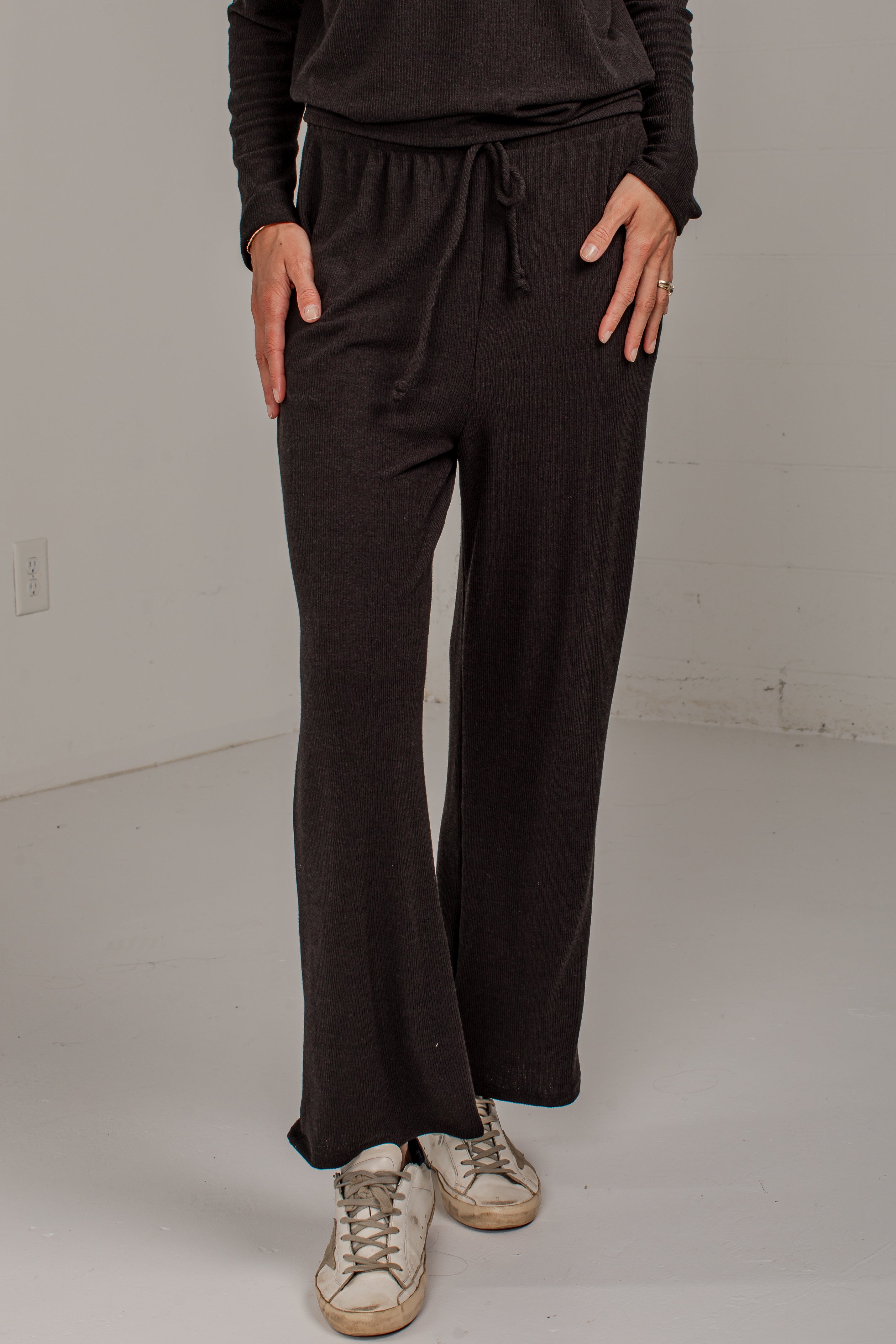 A person wearing dark brown wide-leg drawstring pants with a matching long-sleeve top and distressed white sneakers, standing in a plain studio setting.