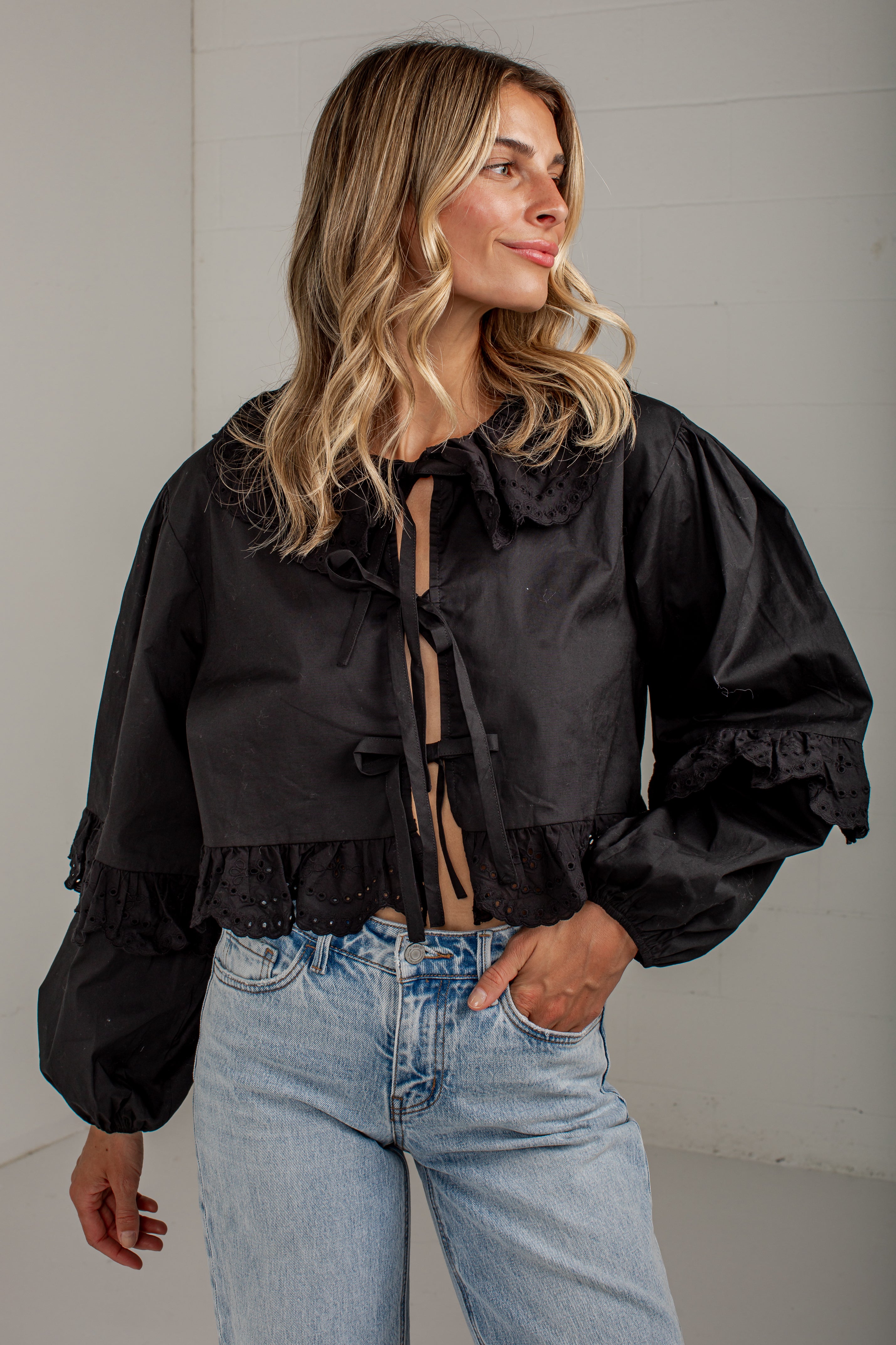 A woman wearing a black cropped satin blouse with dramatic puffed sleeves, ruffled collar and cuffs, and tie-front detailing, paired with light-wash blue jeans against a light gray wall.