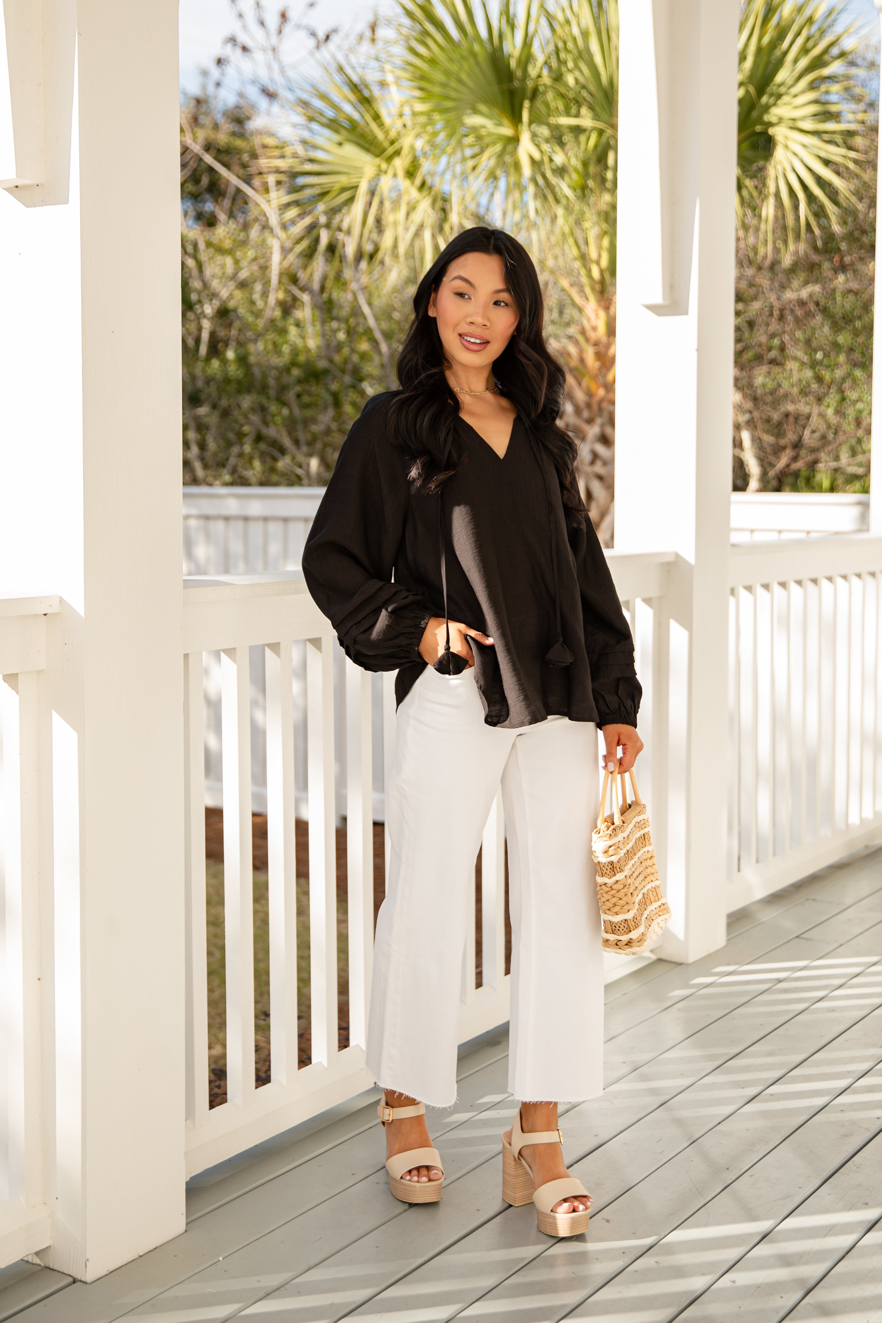 Woman in black blouse and white pants standing on a wooden deck with palm trees in the background