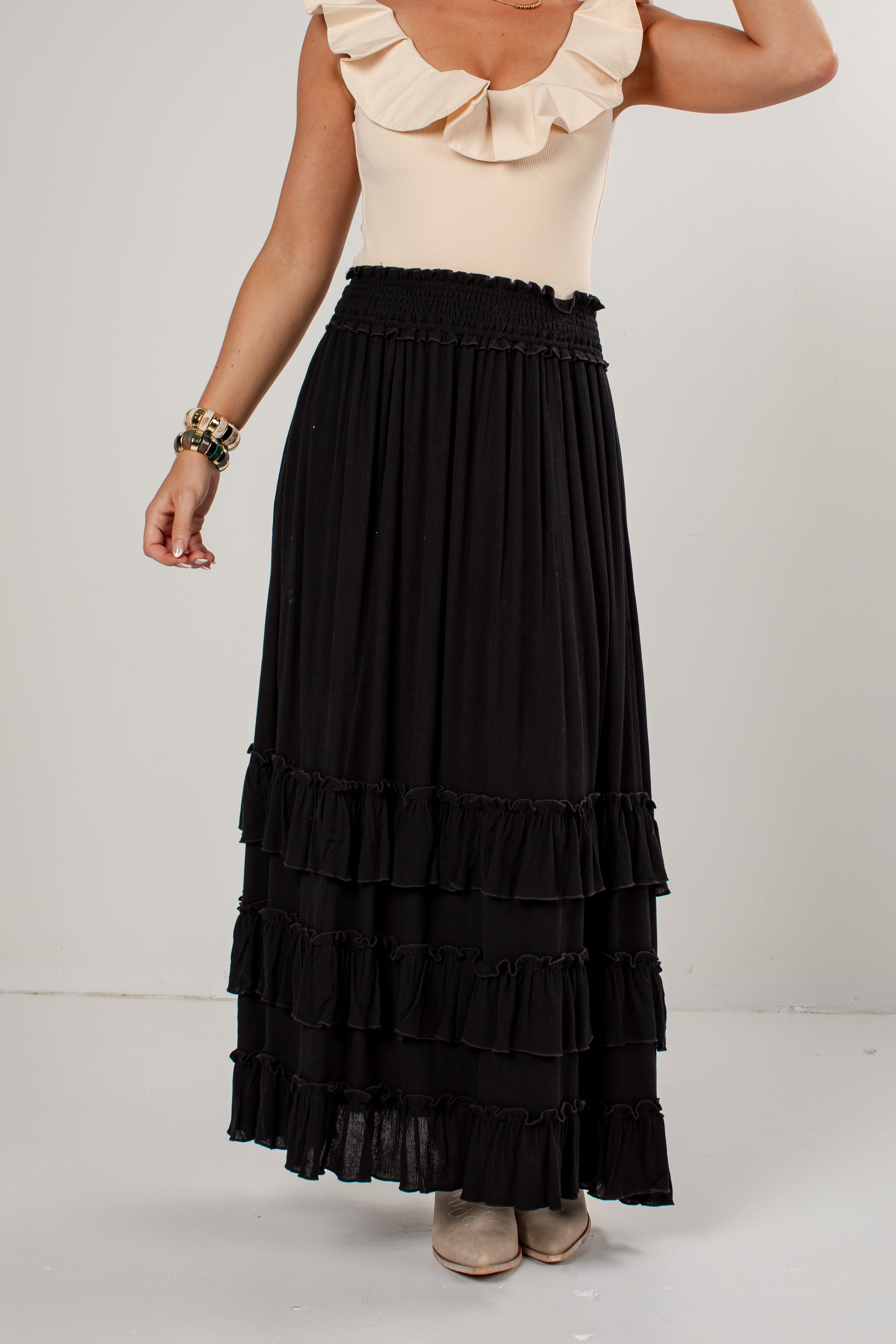 A model wearing a long black tiered maxi skirt with a smocked waistband, paired with a cream ruffled top and beige shoes, shown from the chest down against a plain light gray studio background.