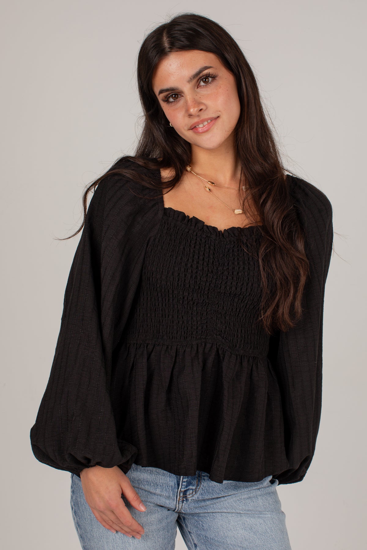 A woman wearing a black smocked long-sleeve blouse with a square neckline and light blue jeans, photographed against a plain light gray studio background.