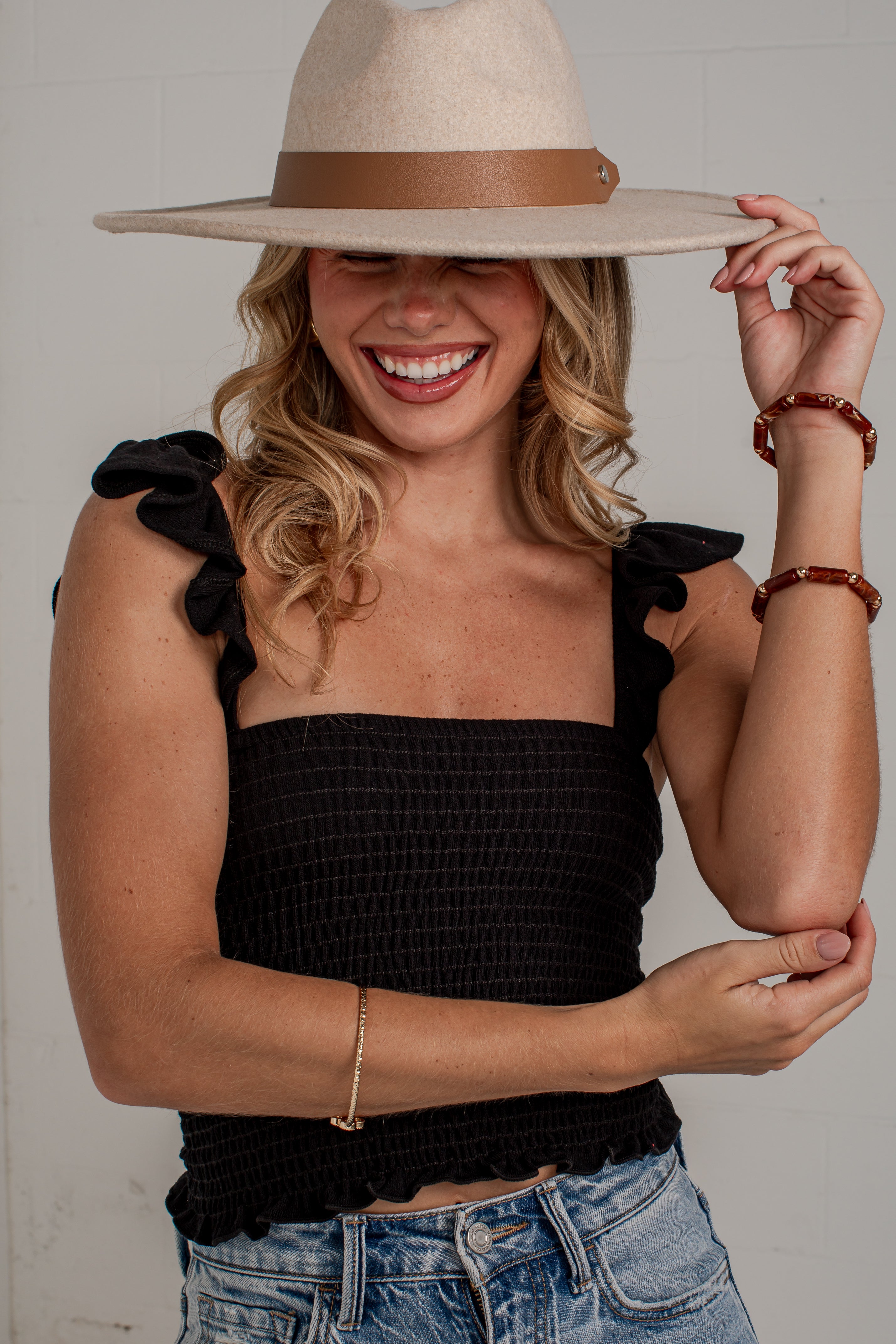 A woman wearing a black smocked crop top with ruffled shoulder straps, blue jeans, and a beige wide-brim hat with a brown band, smiling against a light gray wall.