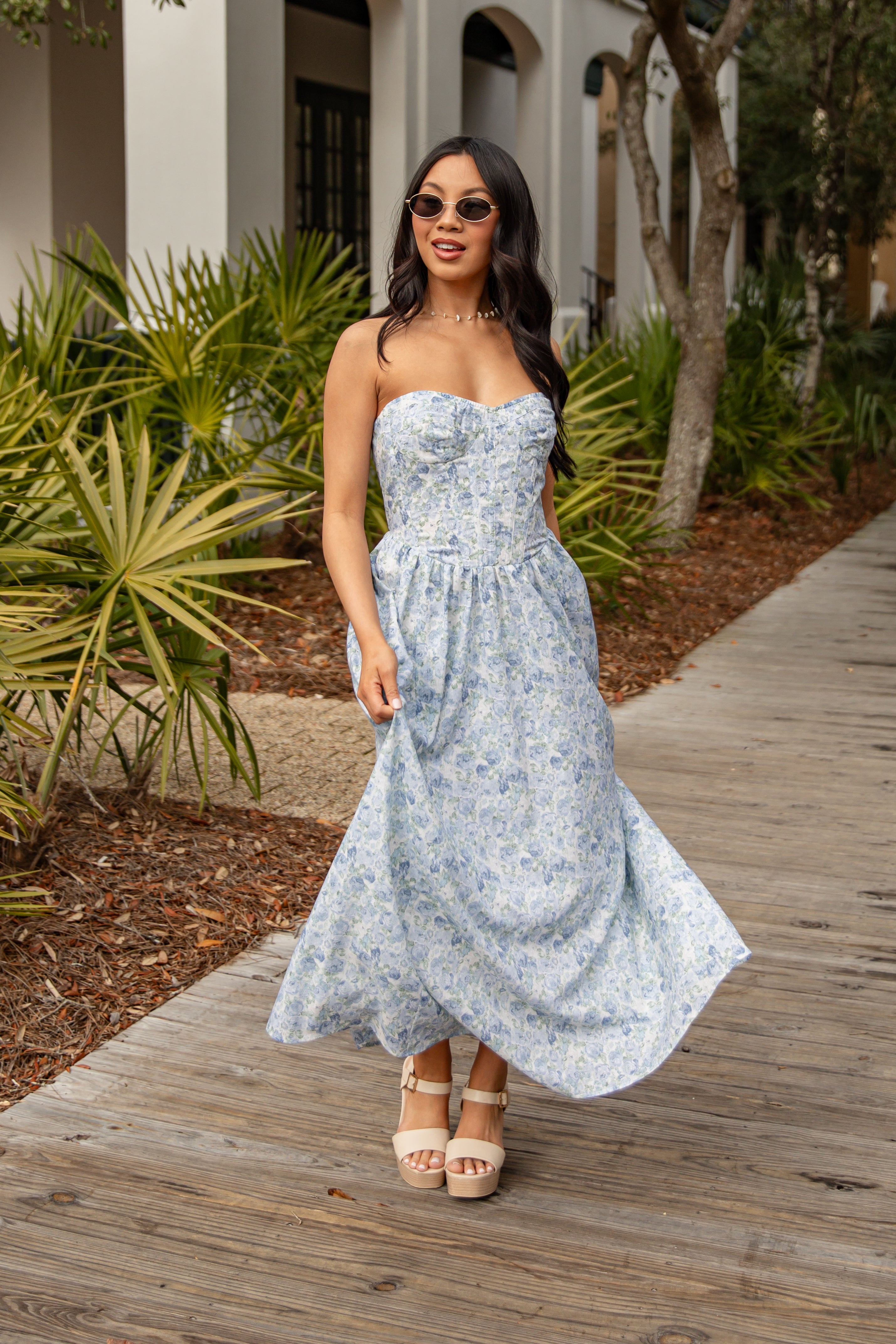 Woman in a strapless floral dress standing on a wooden path with plants and a building in the background.