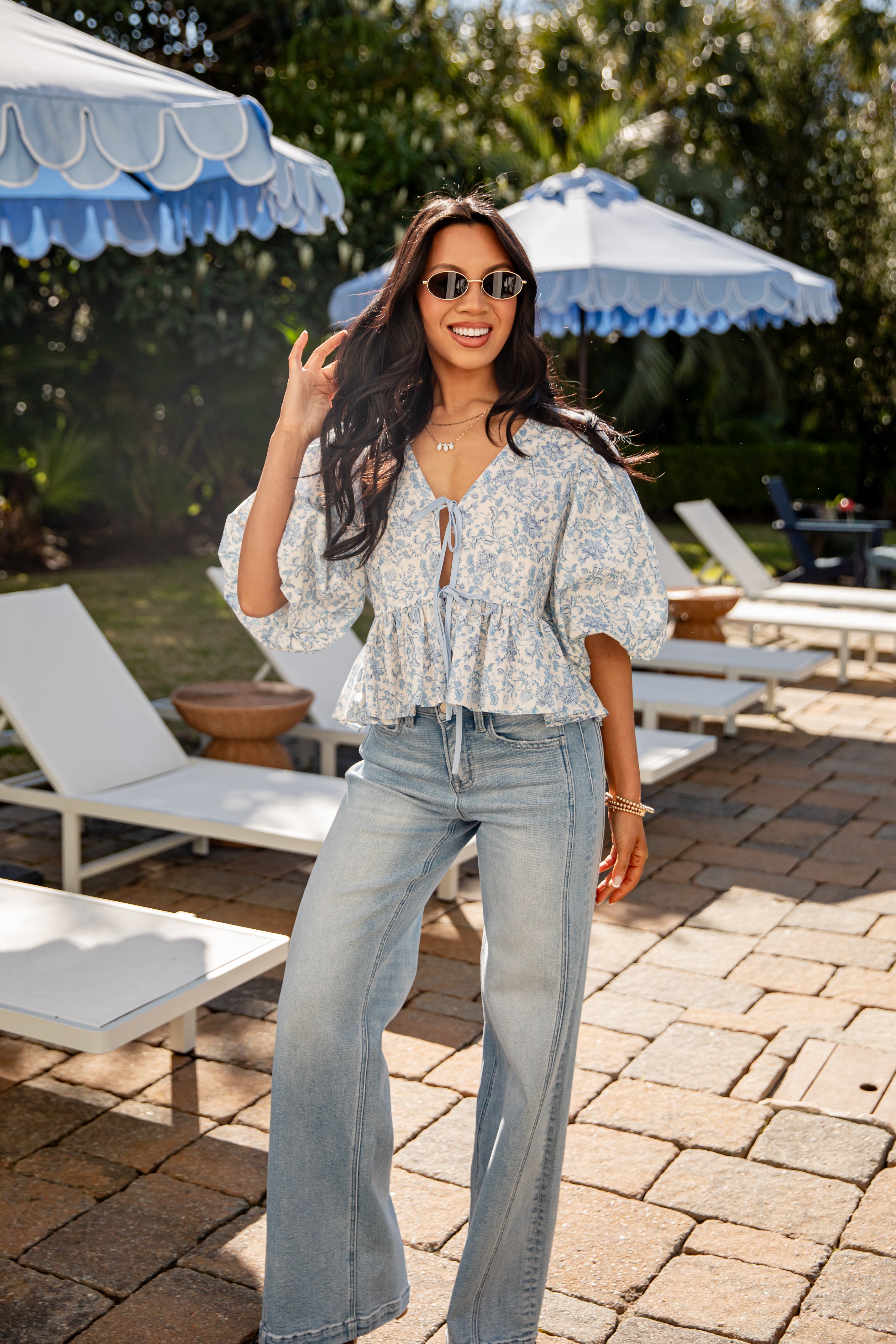 Woman in a floral blouse and jeans standing in a sunlit outdoor setting with lounge chairs and umbrellas.