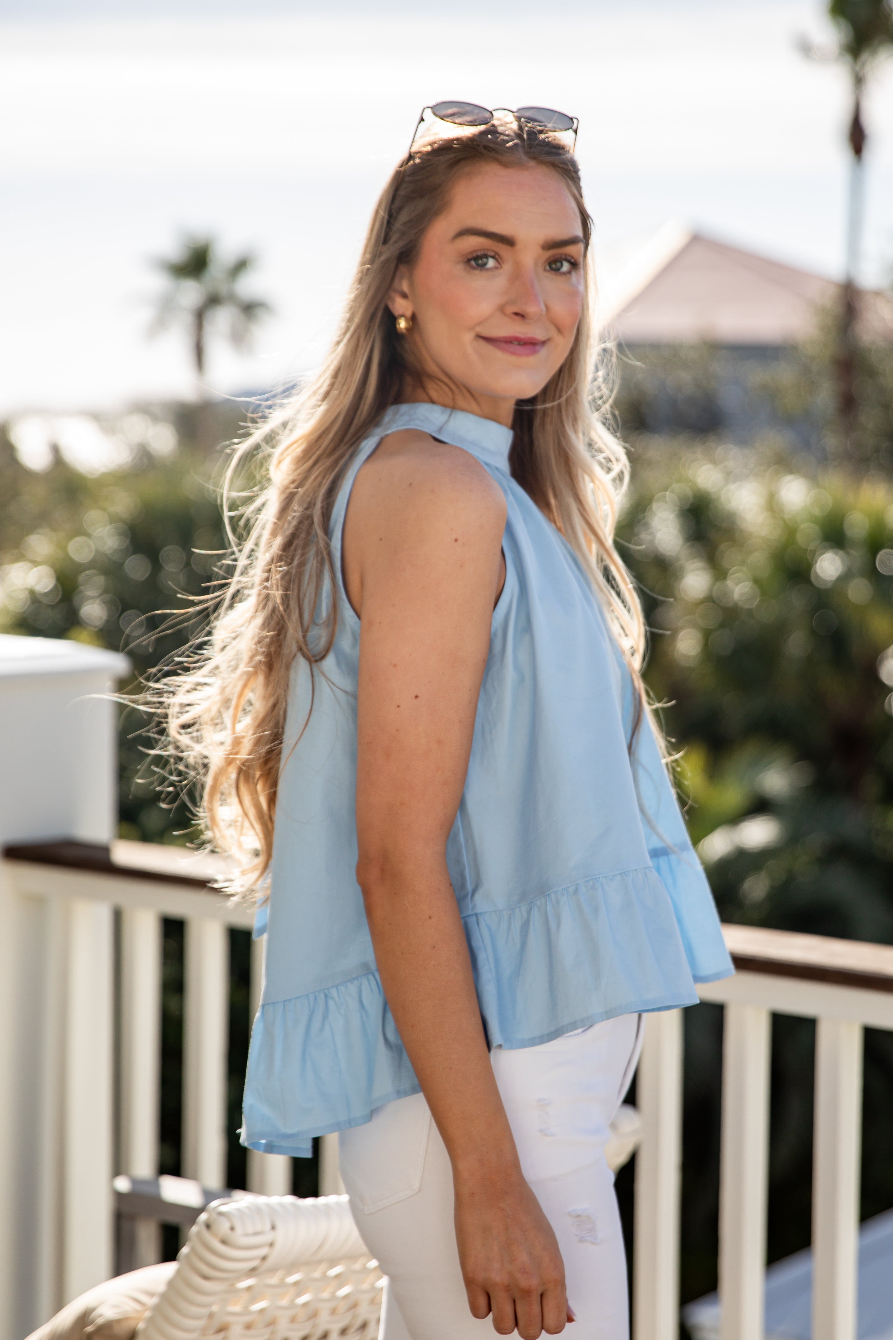 Woman wearing a light blue sleeveless top and white pants on a balcony with greenery in the background.