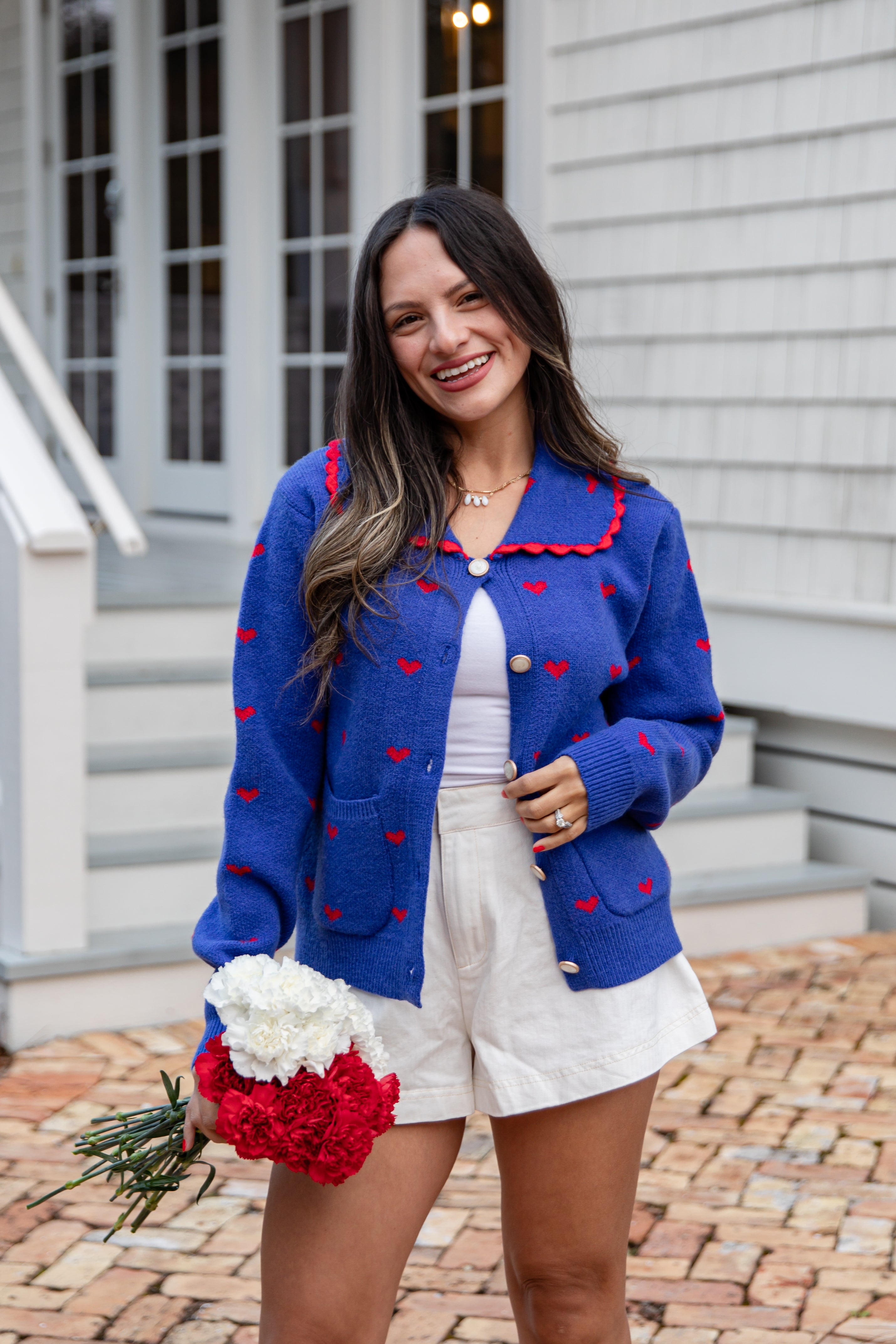 Woman wearing a blue cardigan with red accents, holding flowers, standing outside a house.