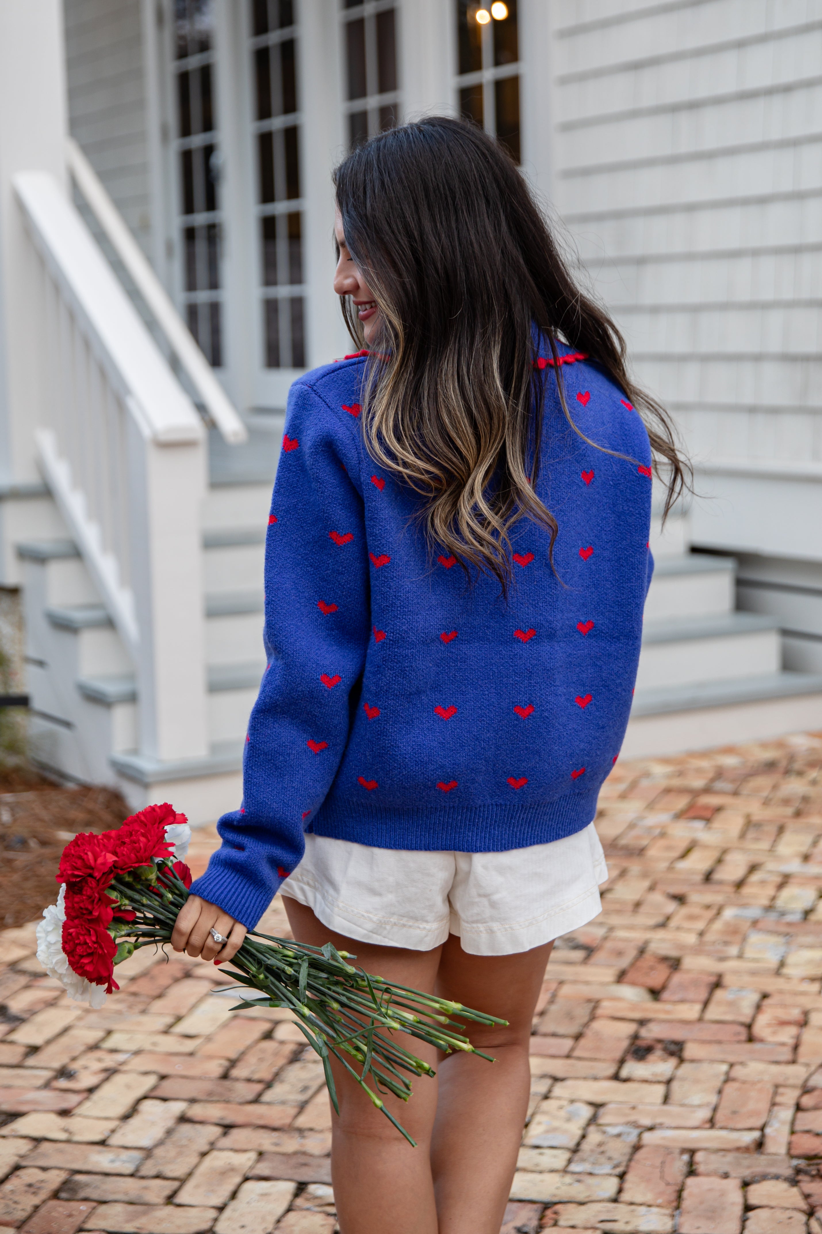 Woman wearing a blue sweater with red heart patterns, holding flowers, standing on a brick path.
