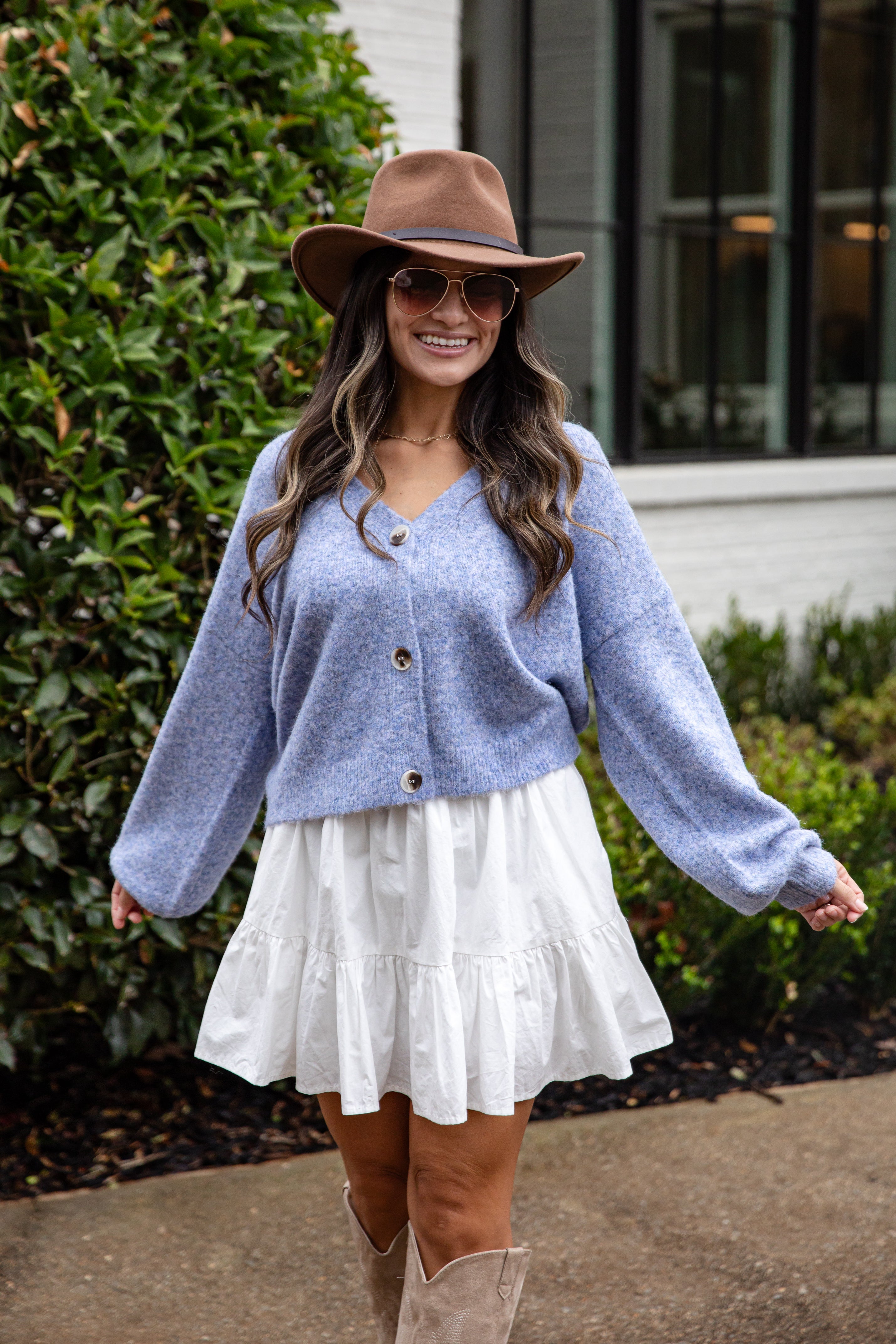 Woman wearing a blue cardigan, white skirt, and brown hat outdoors.