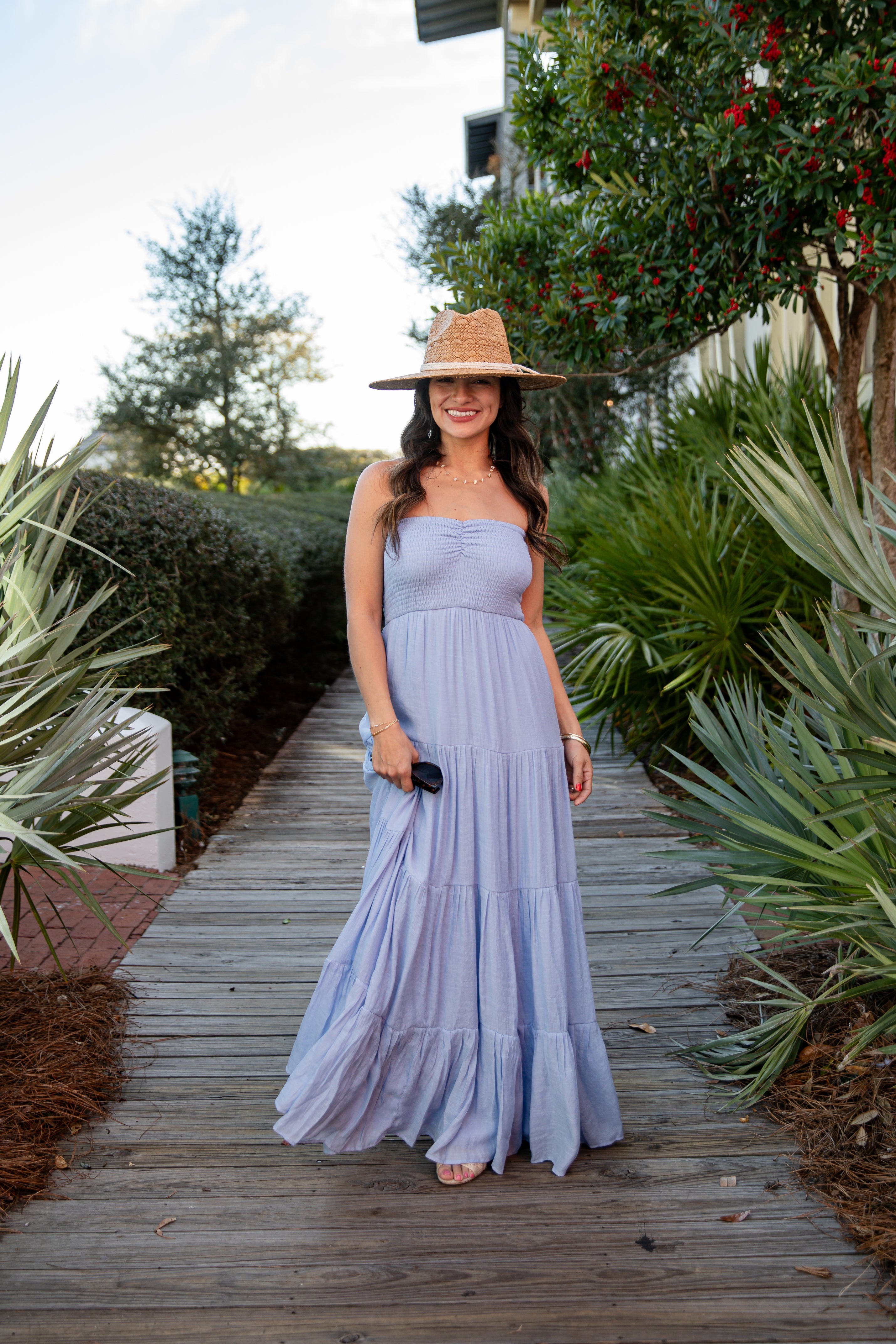 Woman in a purple dress and hat standing on a wooden path with greenery around.