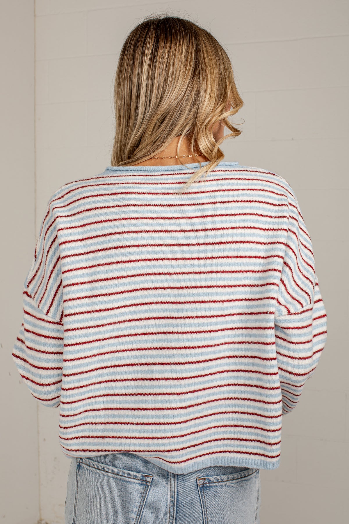 A woman viewed from behind wearing a loose knit sweater with thin horizontal red and white stripes, paired with light blue jeans, against a light neutral wall.