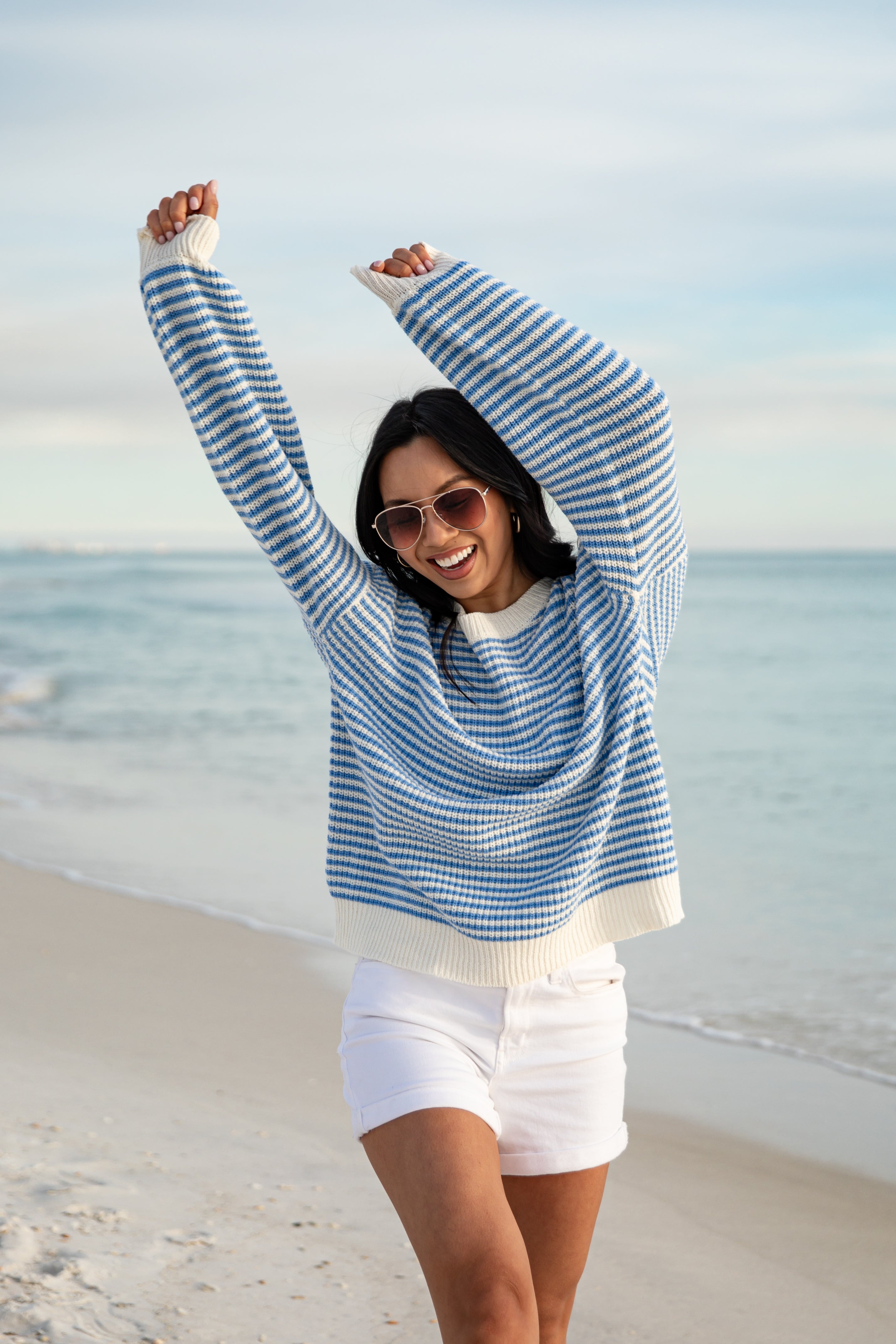 Woman in a blue and white striped sweater and white shorts standing on a beach.