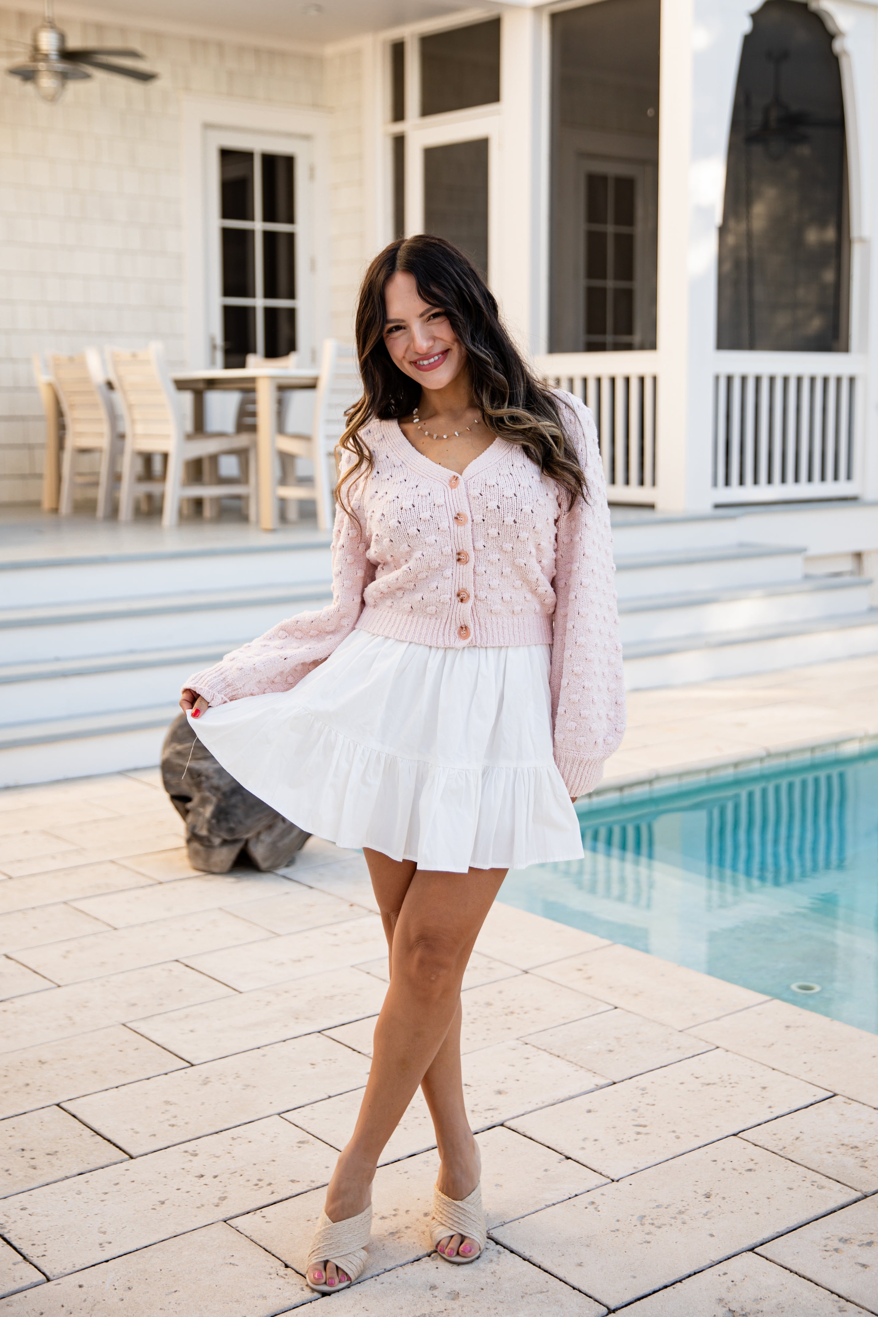 Woman in a pink cardigan and white skirt standing by a poolside.
