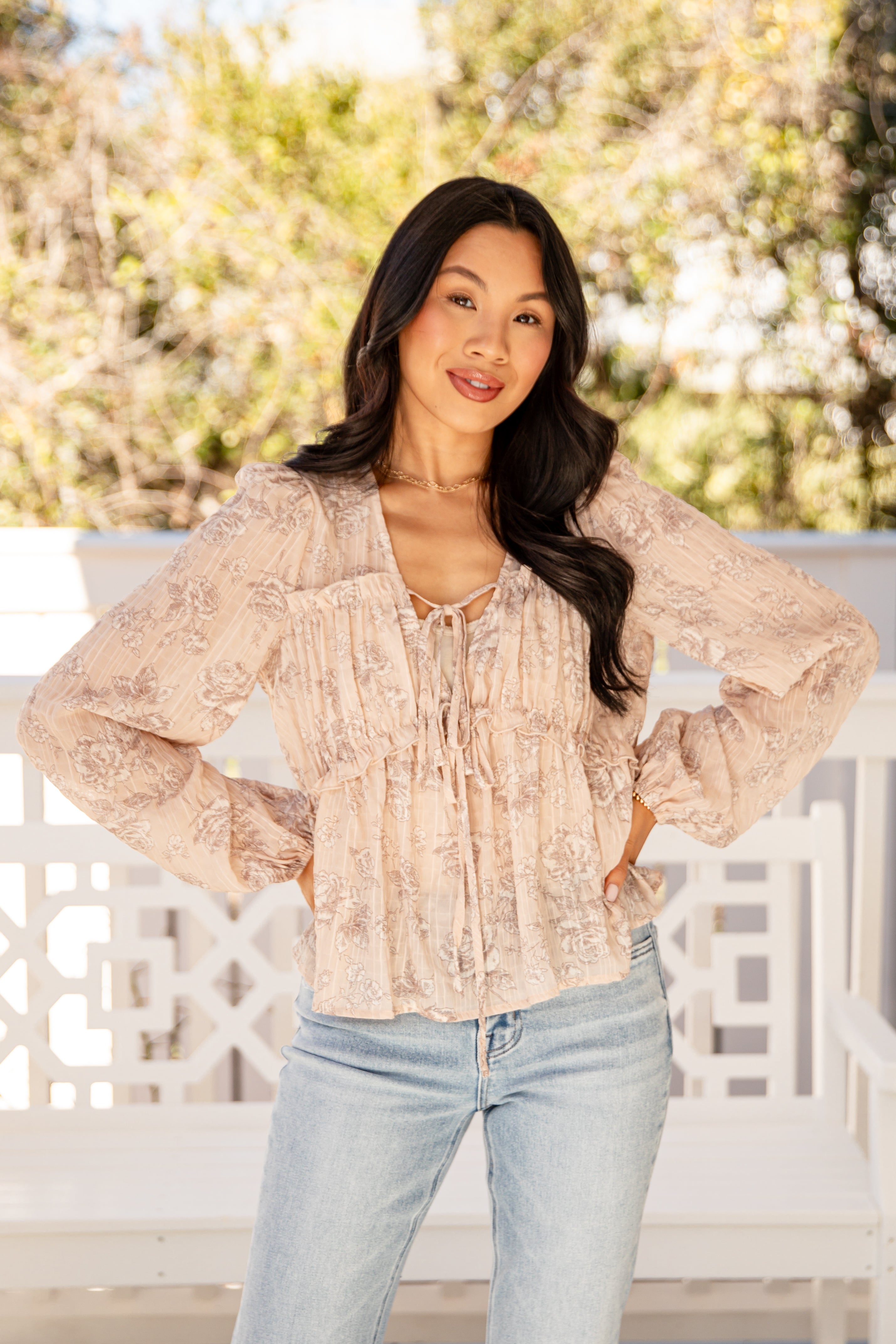Woman wearing a beige blouse and light blue jeans standing outdoors with trees in the background