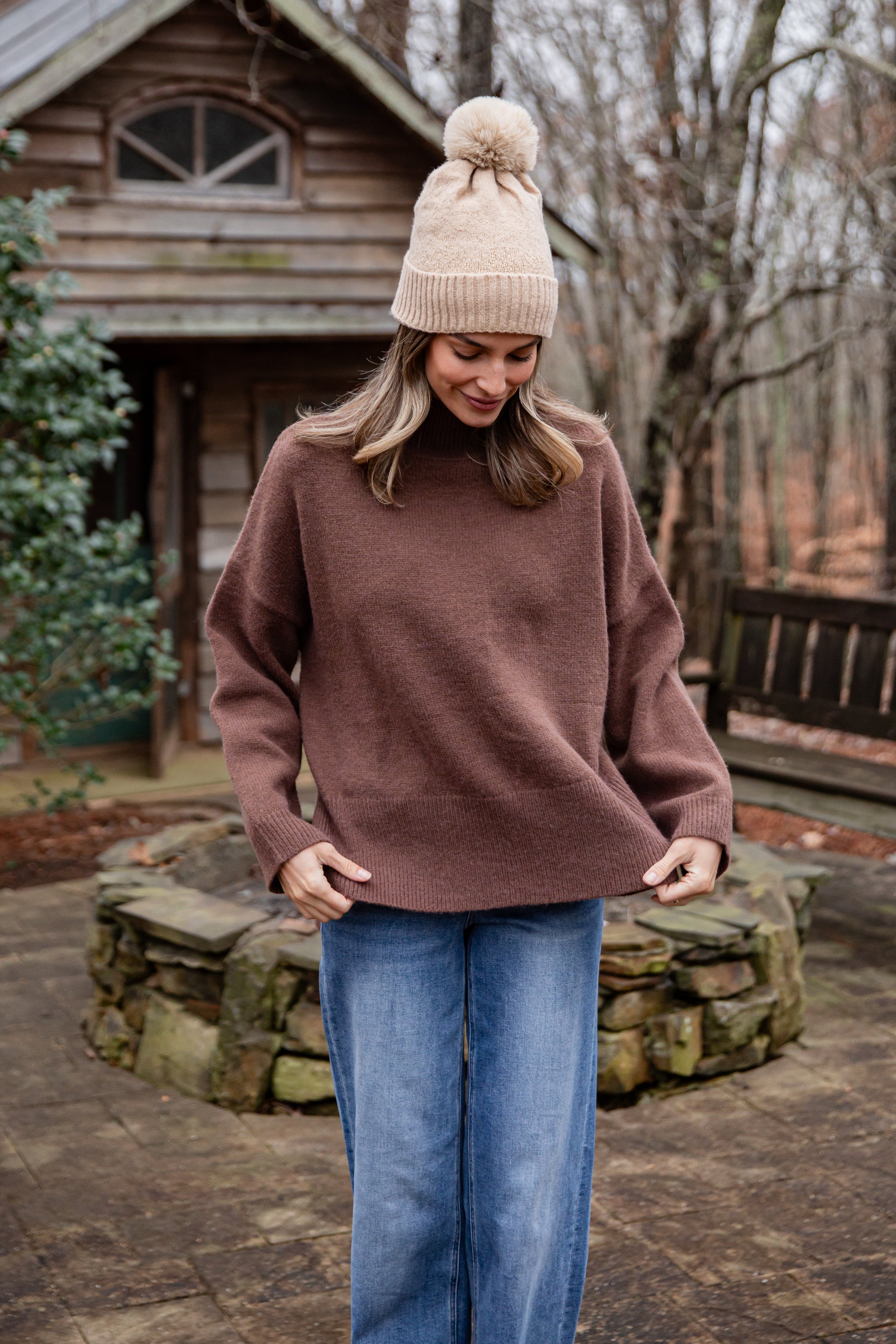 Woman wearing a brown sweater and beige beanie in front of a wooden cabin.