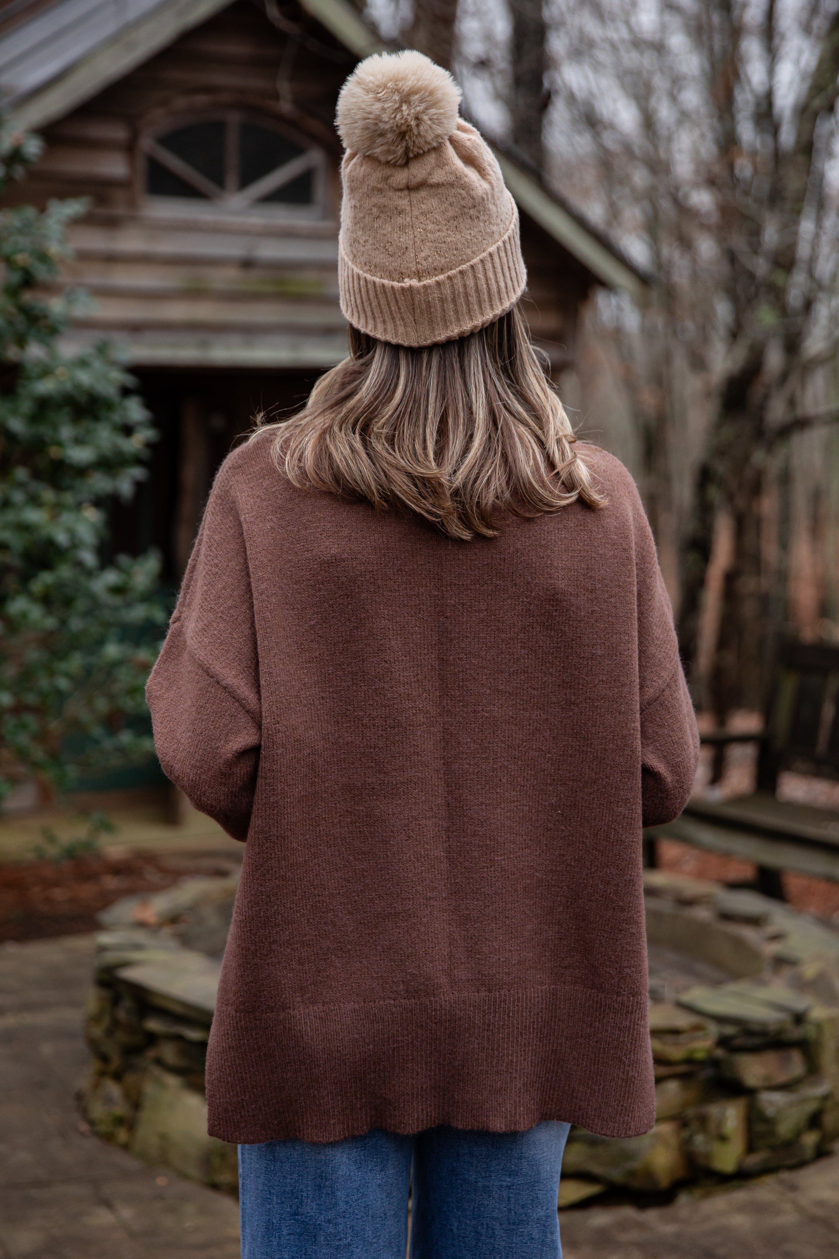 Person wearing a brown coat and beige beanie with pom-pom, standing in front of a wooden cabin.