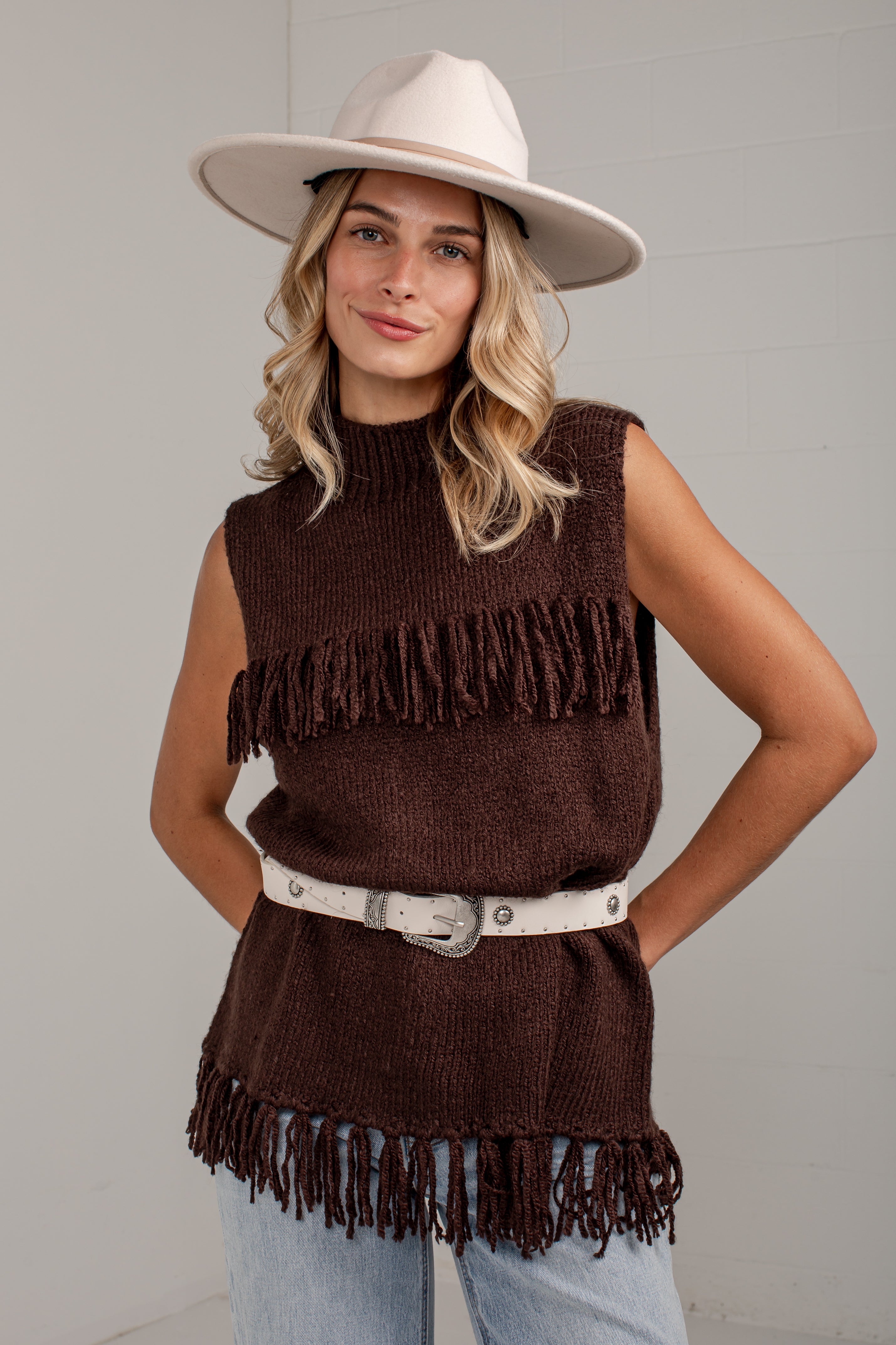 A woman modeling a sleeveless brown knit top with fringe trim, styled with a white belt, light blue jeans, and a cream wide-brim hat against a light gray wall.