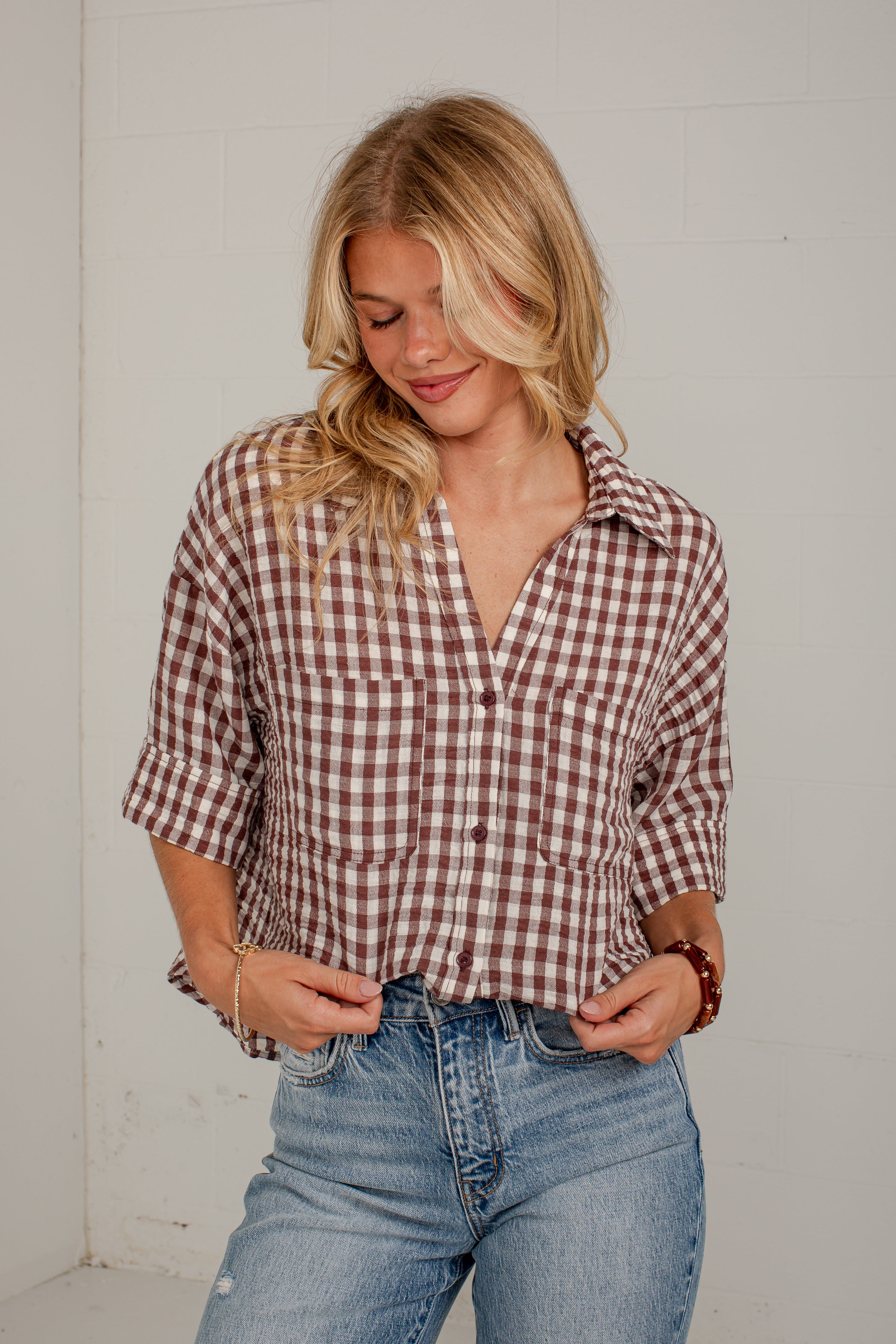 A woman wearing a brown and white gingham plaid button-down shirt with three-quarter sleeves and blue jeans, standing against a white wall.