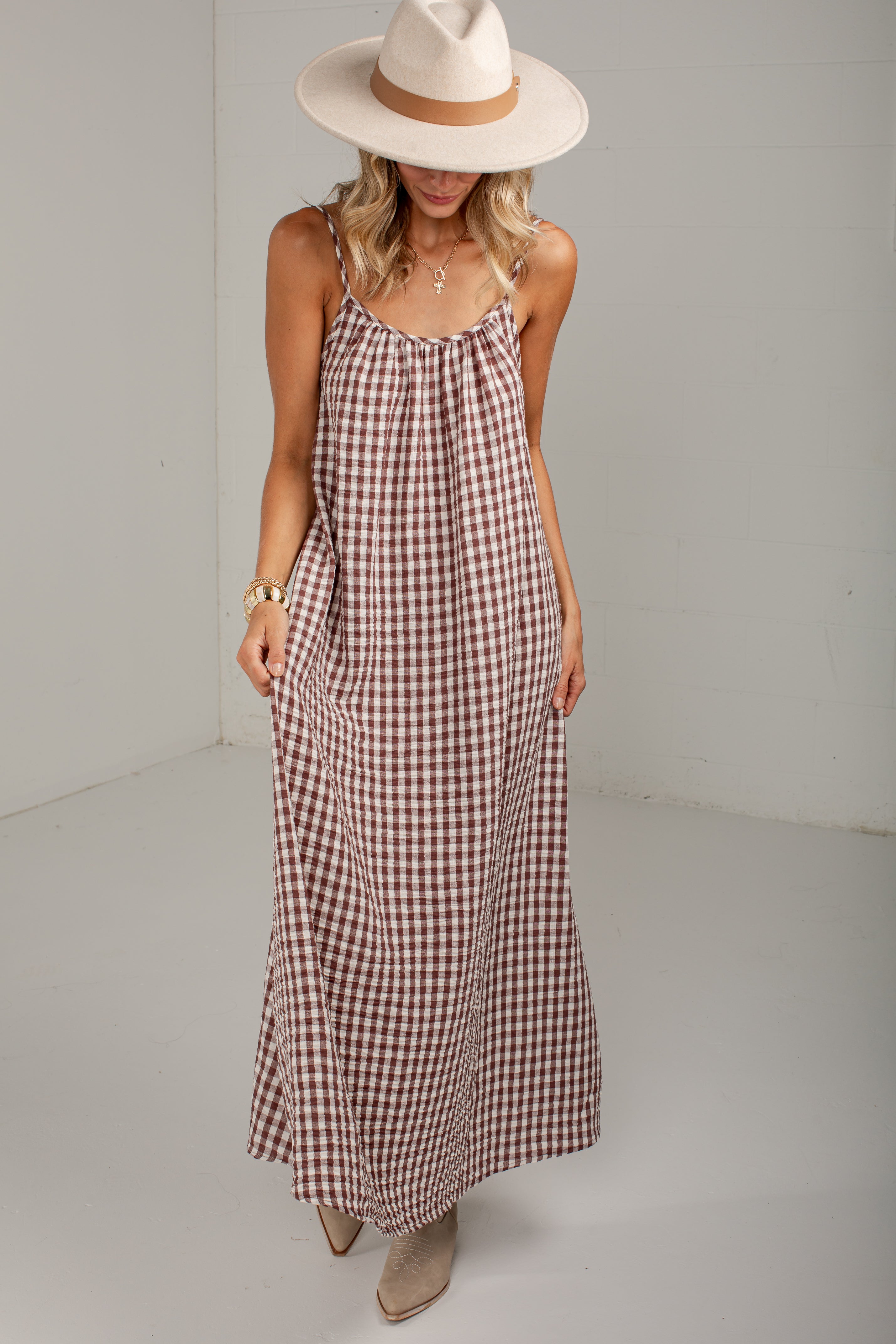A woman modeling a brown and beige gingham maxi dress with spaghetti straps, styled with a wide-brimmed cream hat and beige ankle boots.