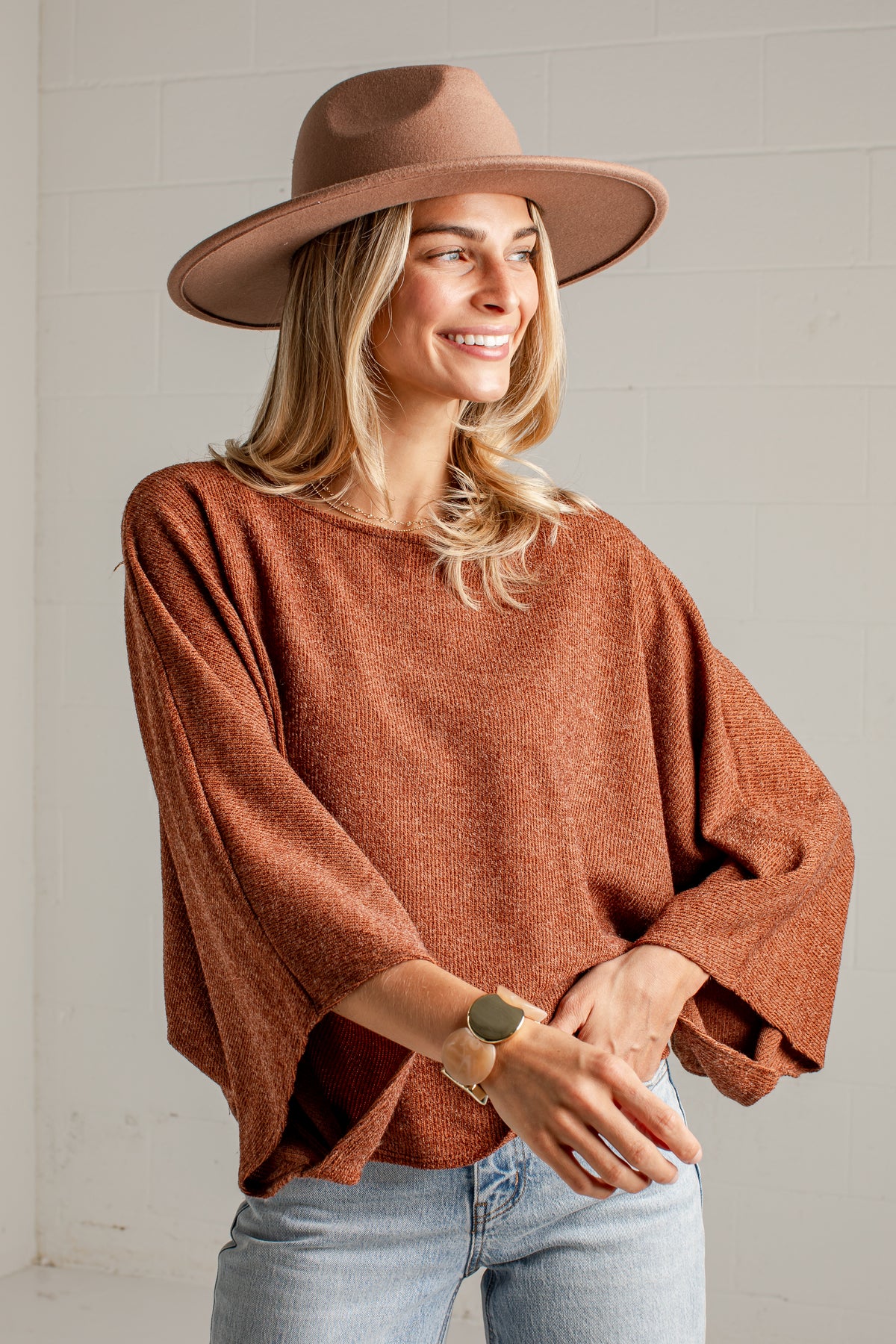 A woman wearing a rust-brown knit sweater, a wide-brimmed brown hat, and light-wash blue jeans, standing against a white brick wall.