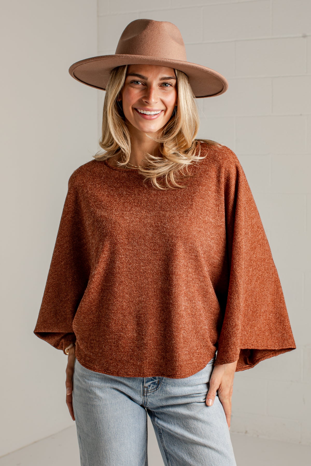 A woman wearing a rust-brown knit sweater, a wide-brimmed brown hat, and light-wash blue jeans, standing against a white brick wall.