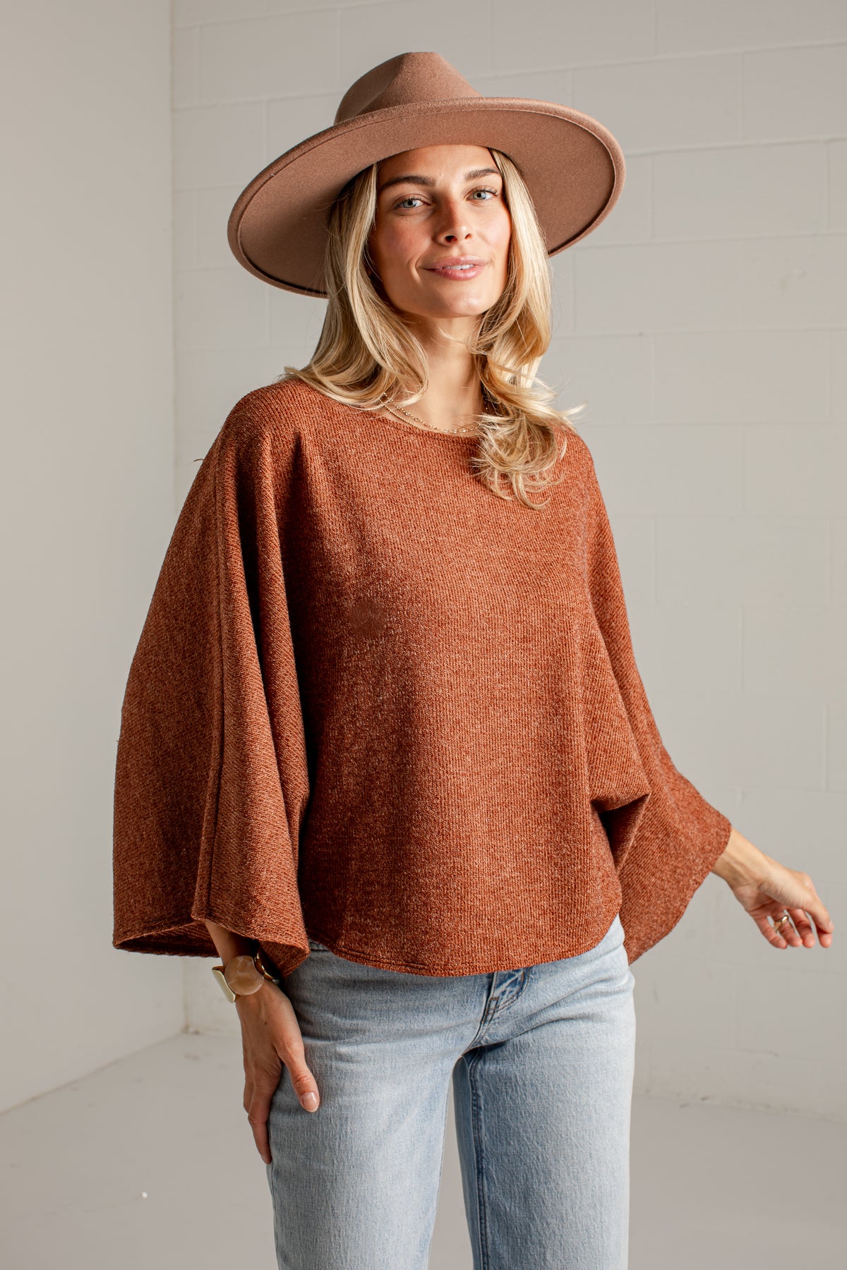 A woman wearing a rust-brown knit poncho sweater, a wide-brimmed brown hat, and light-wash blue jeans, standing against a light gray wall.