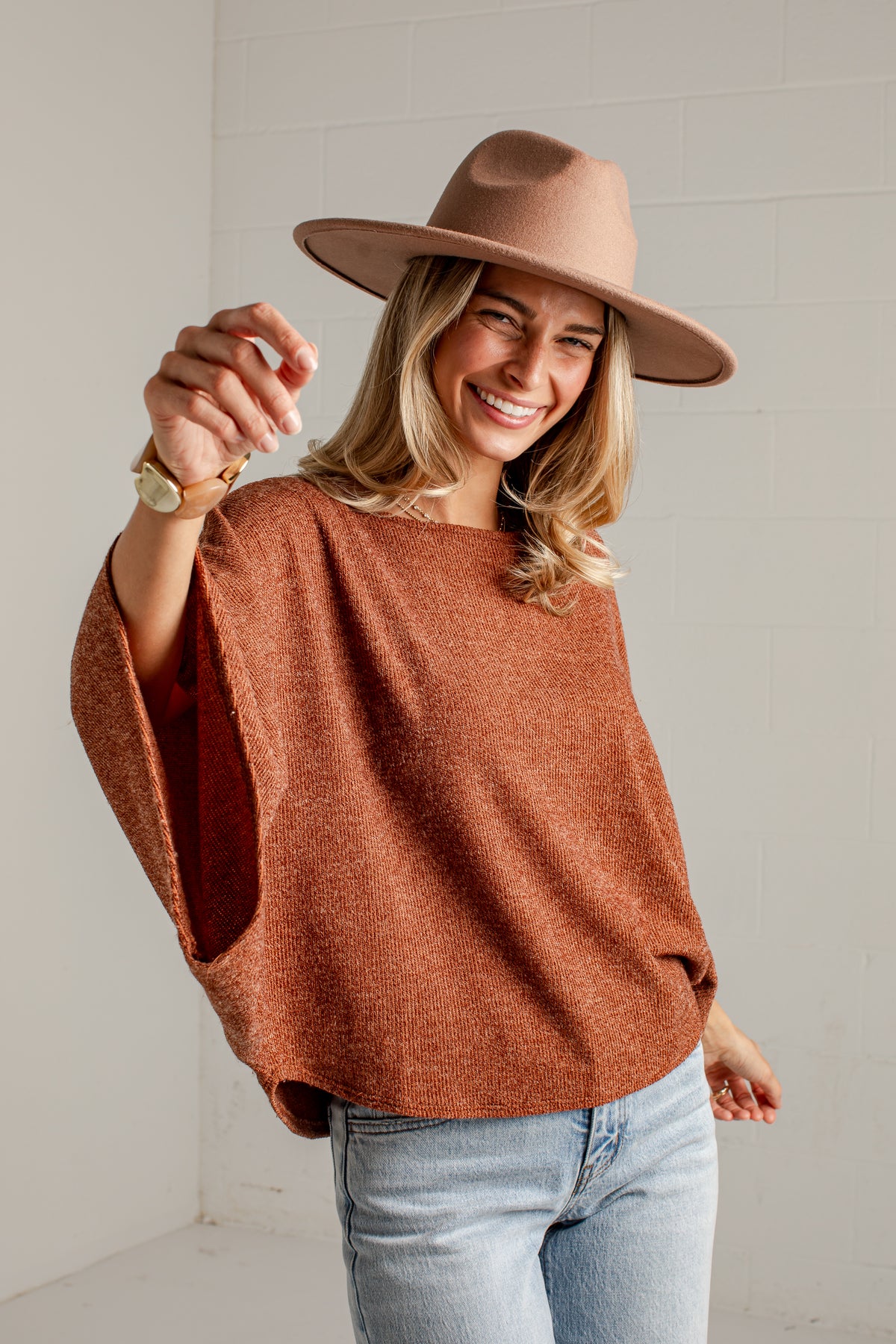 A woman wearing a rust-orange knit poncho sweater, a brown wide-brim hat, and light-wash blue jeans, smiling and pointing toward the camera in a bright indoor setting.