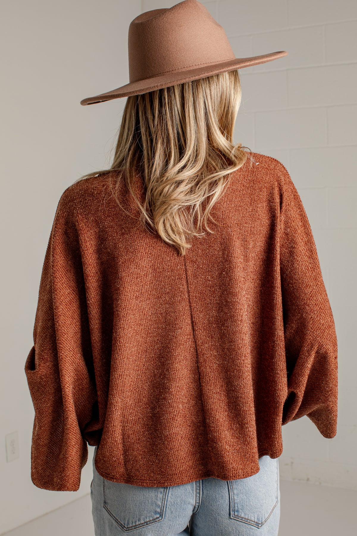 A woman viewed from behind wearing a rust-brown knit poncho sweater, a brown wide-brim hat, and light-wash blue jeans against a light gray wall.