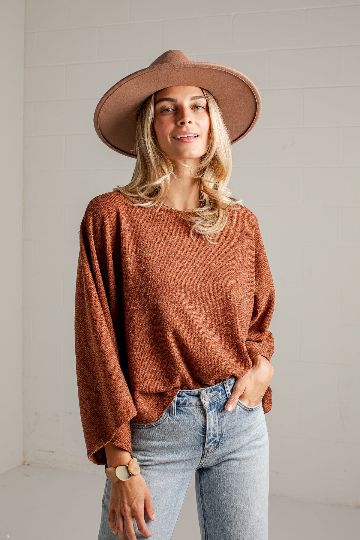 A woman wearing a rust-brown knit sweater, a wide-brimmed brown hat, and light-wash blue jeans, standing against a white brick wall.
