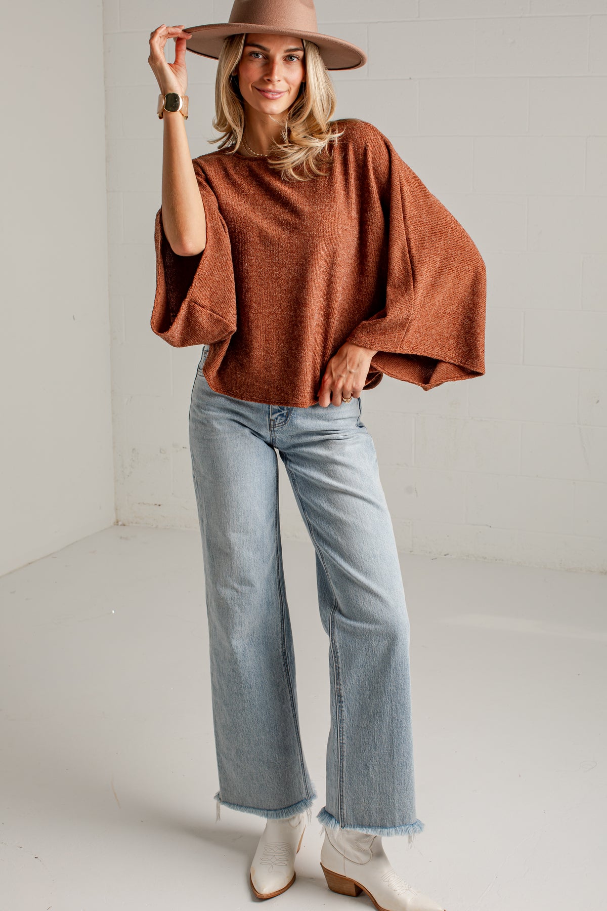 A woman wearing a rust-brown knit sweater, a wide-brimmed brown hat, and light-wash blue jeans, standing against a white brick wall.