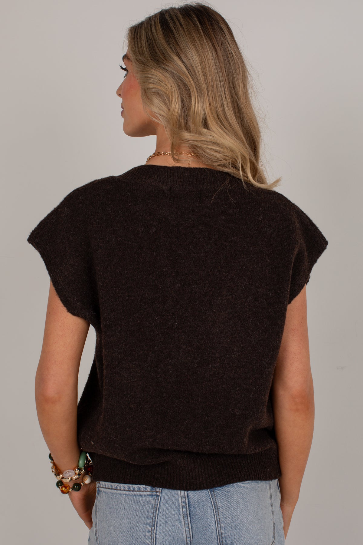 A woman shown from the back wearing a dark brown short-sleeve knit top and light blue jeans against a plain light gray background.