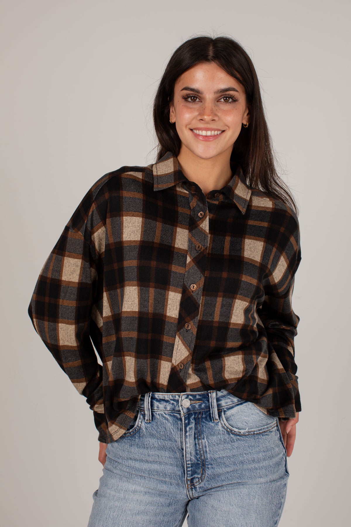 A woman wearing a brown and beige plaid button-down shirt with light blue jeans, standing against a solid light gray background.