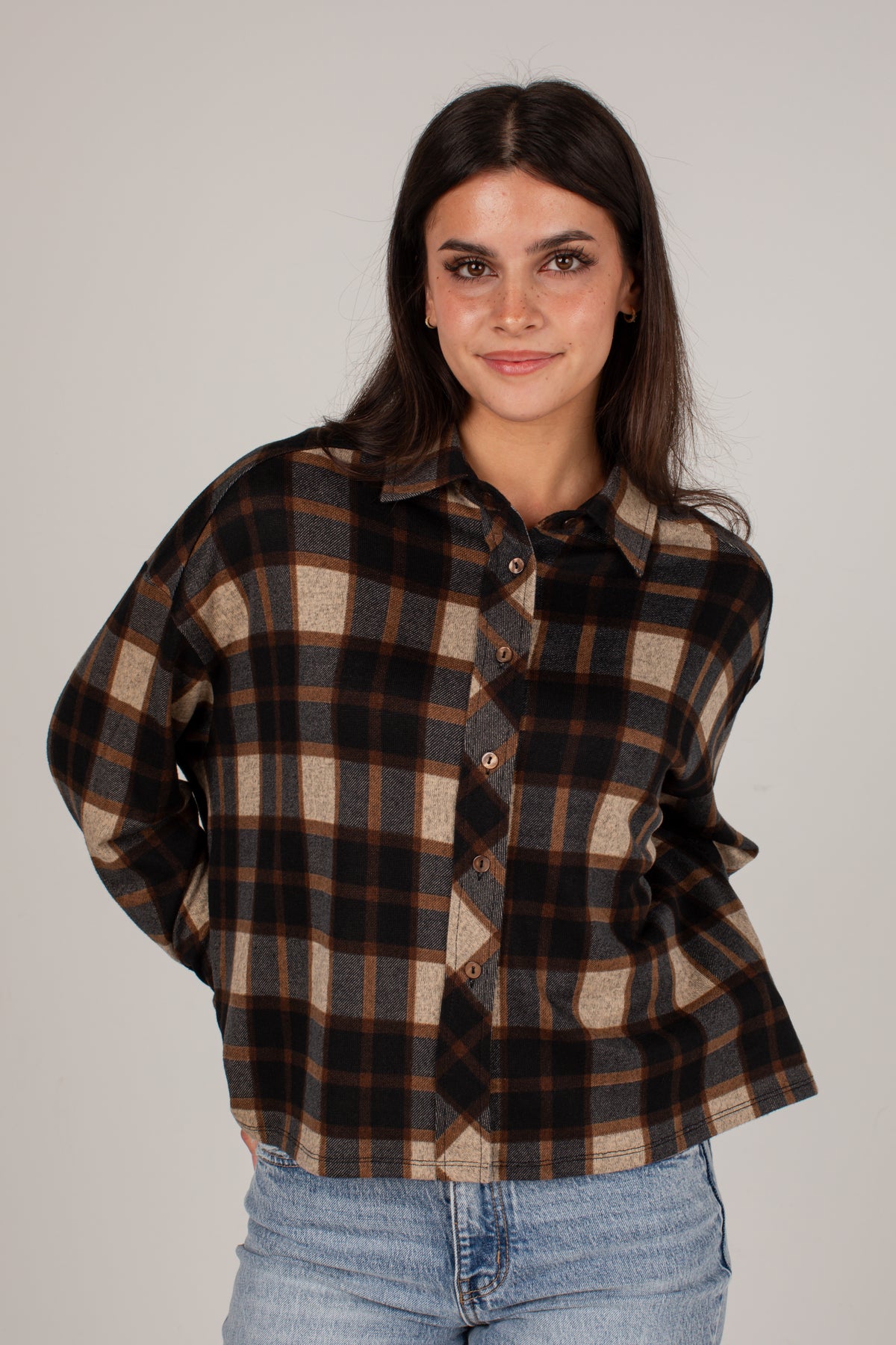 A woman wearing a brown and beige plaid button-up shirt with light blue jeans, standing against a solid light gray background.