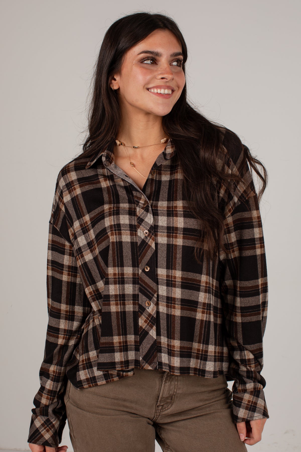 A woman wearing a brown, black, and beige plaid button-up shirt with brown pants, standing against a plain light gray studio background.
