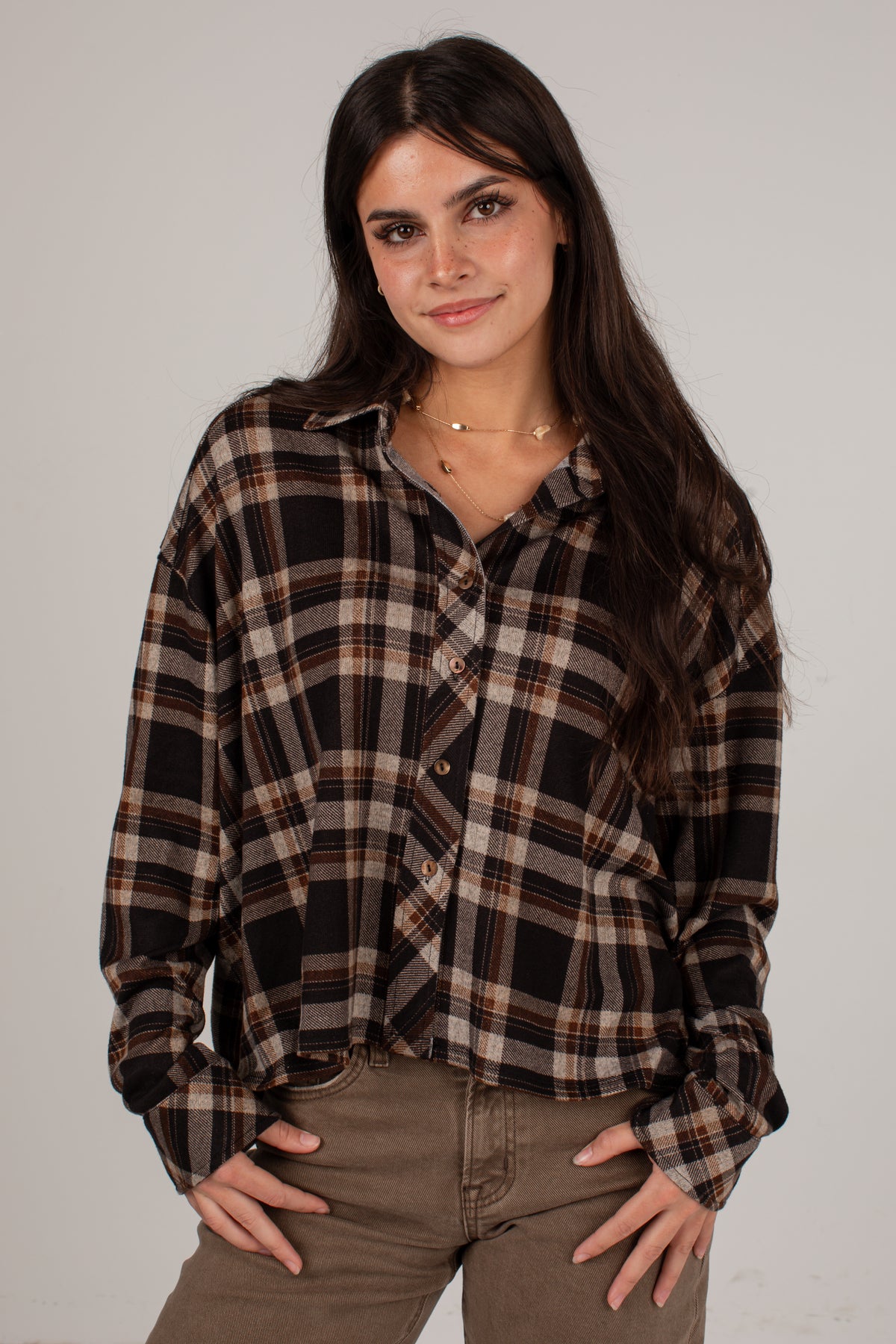 A woman modeling a long-sleeve button-up plaid shirt in black, brown, and beige, paired with brown pants, against a plain light gray studio background.