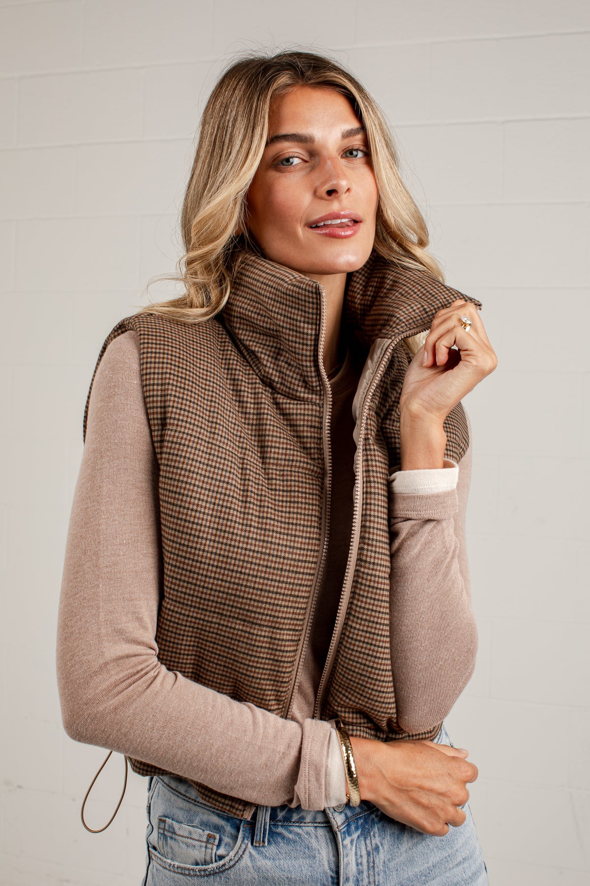 A woman wearing a brown and beige houndstooth zip-front vest over a dusty pink long-sleeve top and blue jeans, standing against a light gray paneled wall.